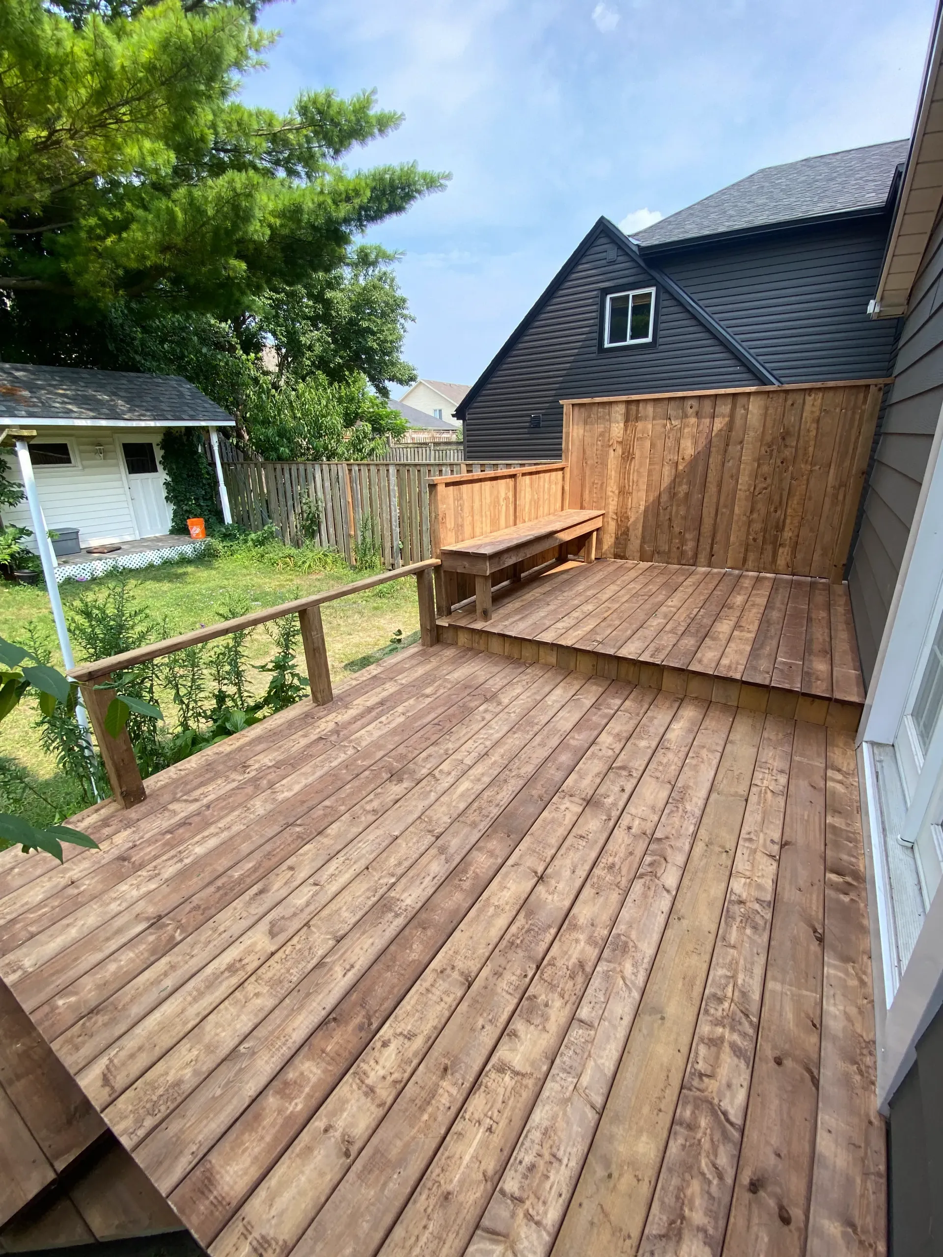 Wooden deck with built-in bench and fence, attached to a gray house. Green trees and a white shed are in the background.
