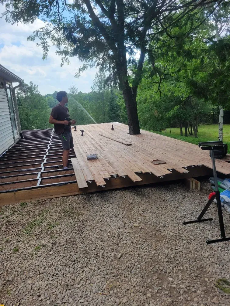 Person standing on a partially built wooden deck, near a house and trees. Gravel ground.