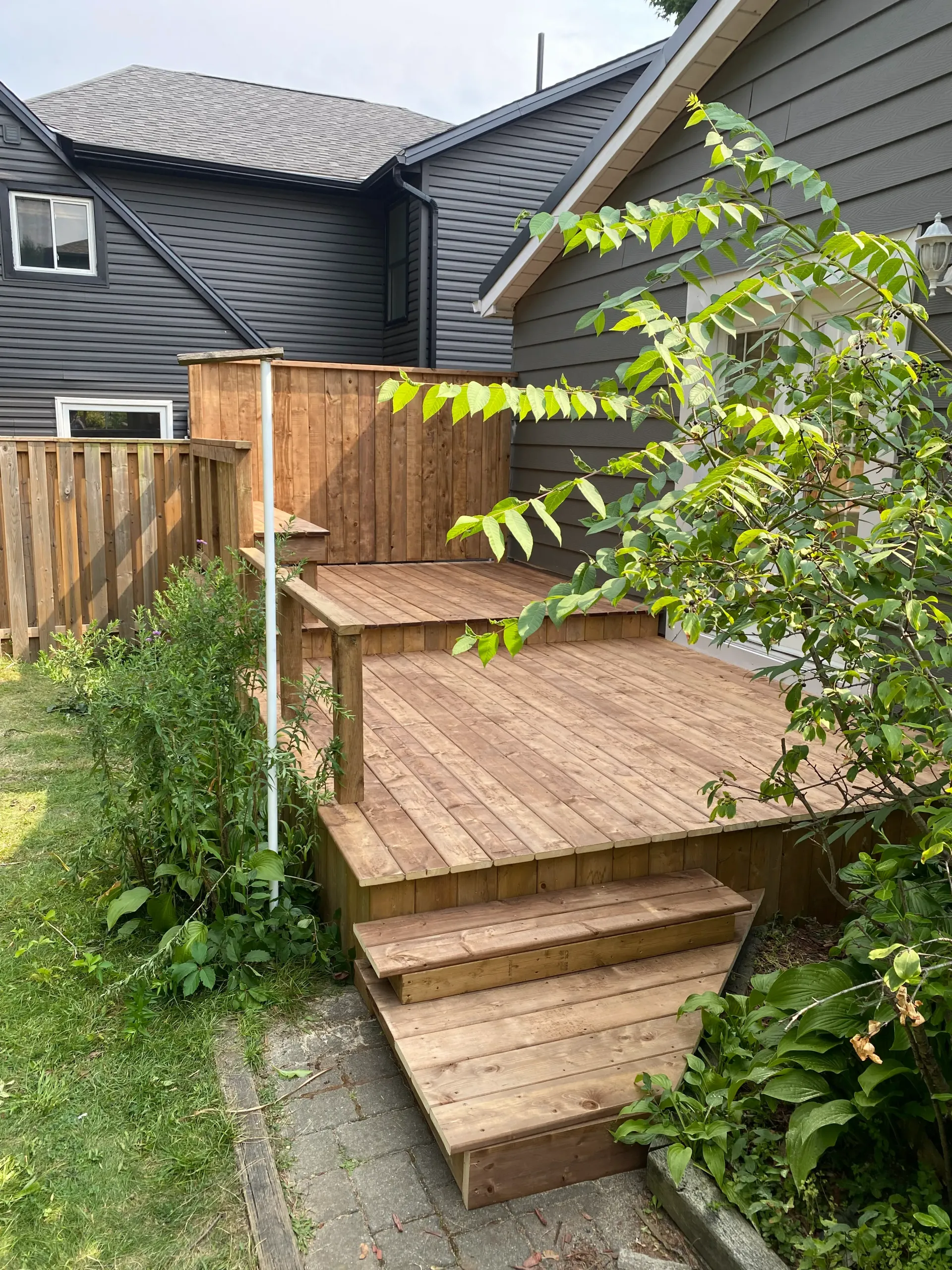 Wooden deck with steps next to a house with dark gray siding. Green foliage surrounds the deck.
