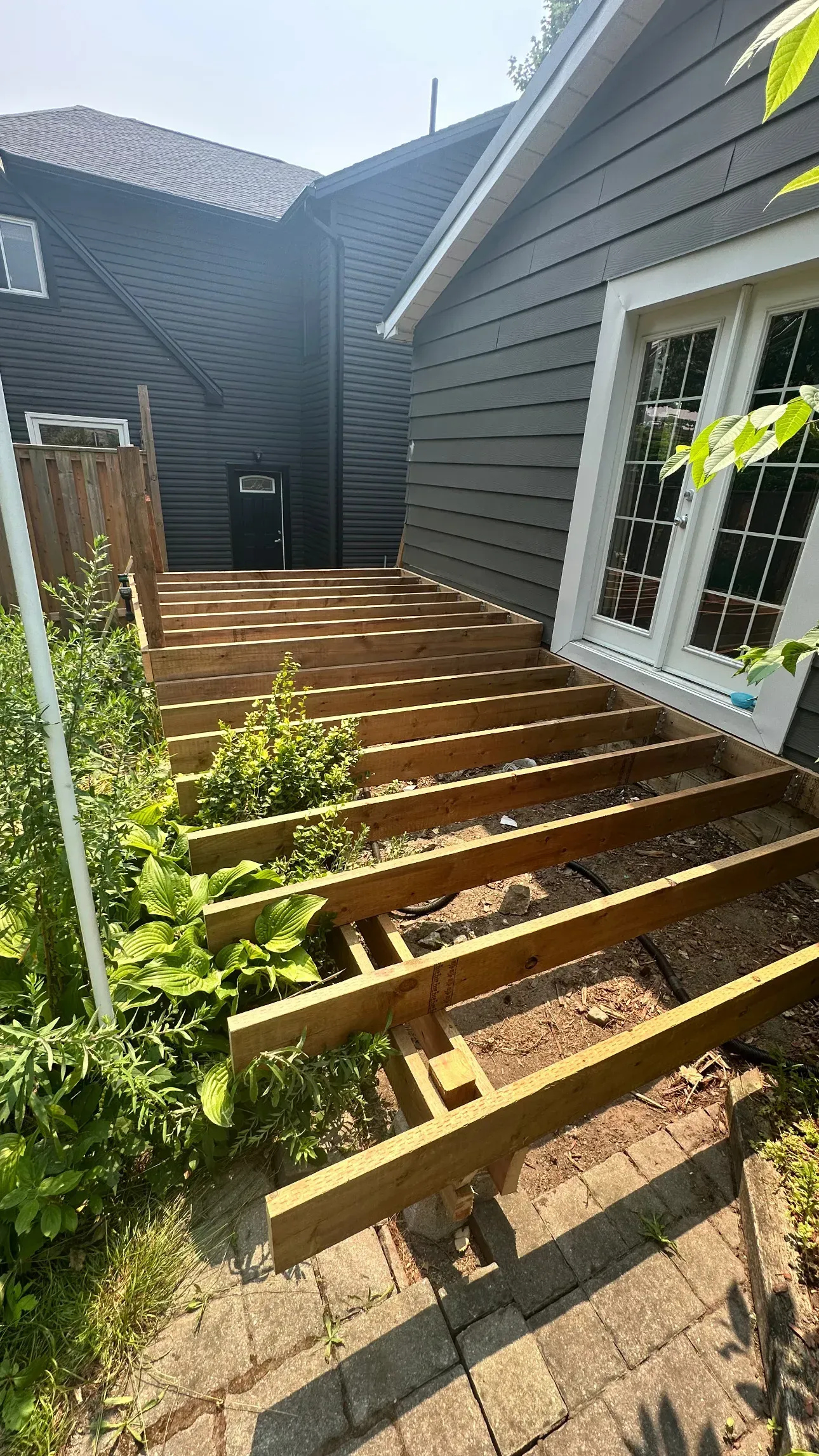 Partially constructed wooden deck next to a dark gray house. Deck has exposed joists, with greenery around.