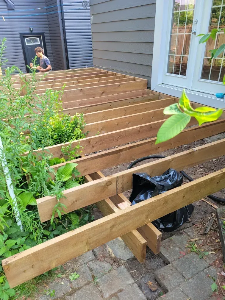 Wooden deck frame under construction next to a house with a person in the background.