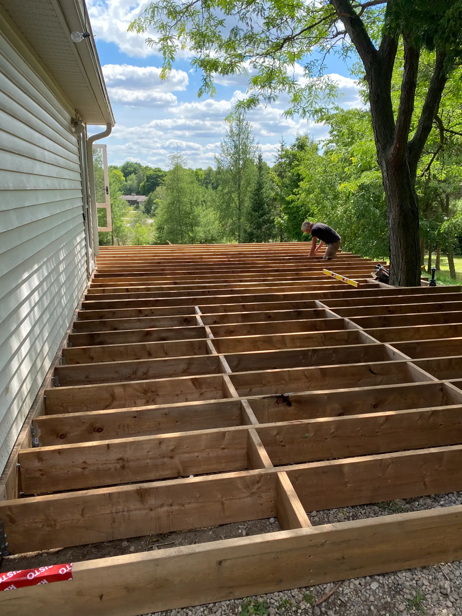 Framed wooden deck under construction next to a house; person working in the background under a blue sky.