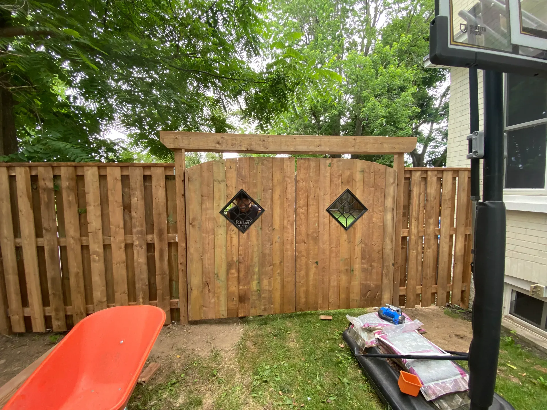 Wooden fence with double gate, diamond-shaped windows, and a pergola top in a backyard setting.