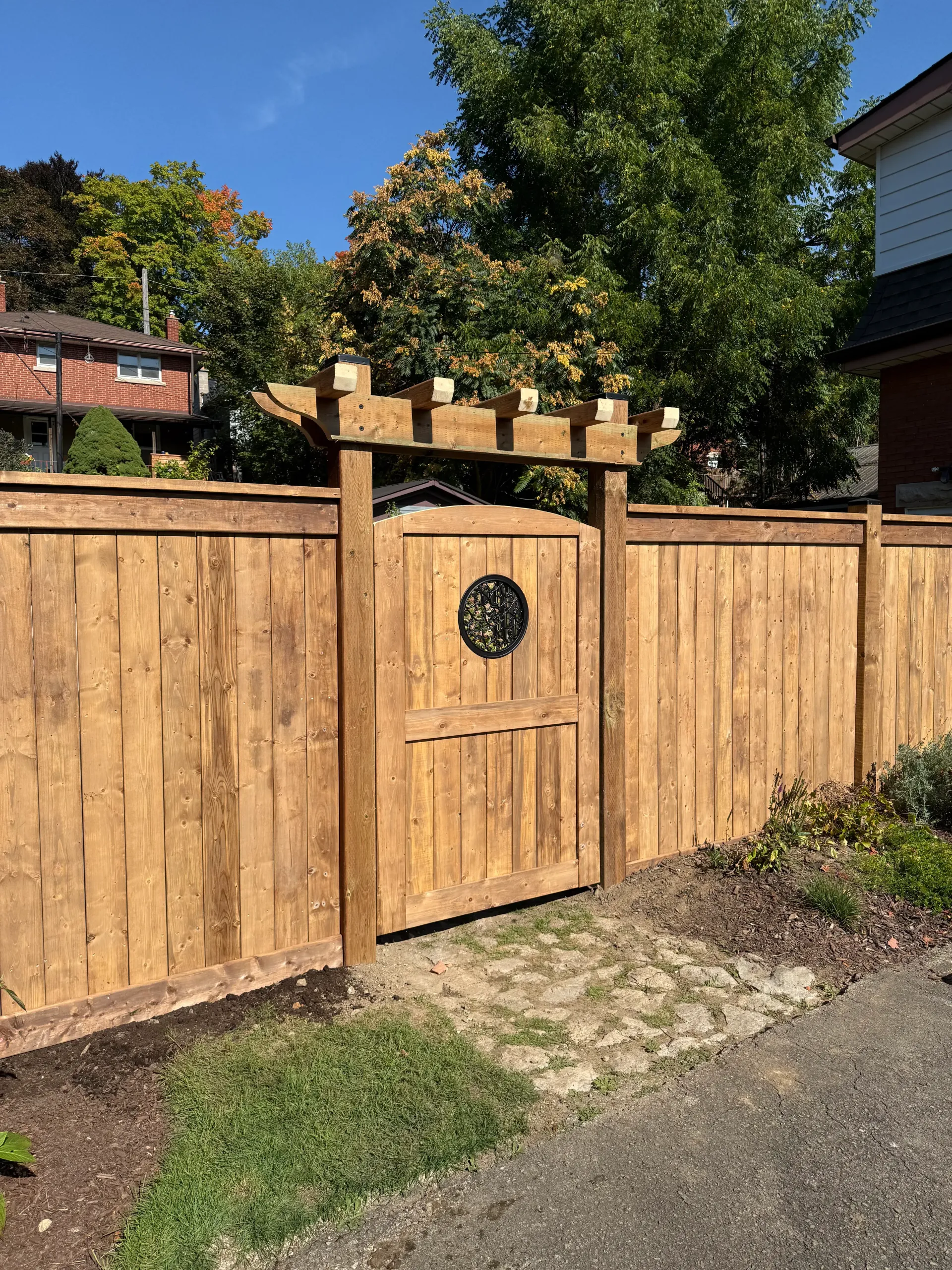 Wooden fence and gate with stone pathway, topped by a decorative arbor; trees and houses in the background.