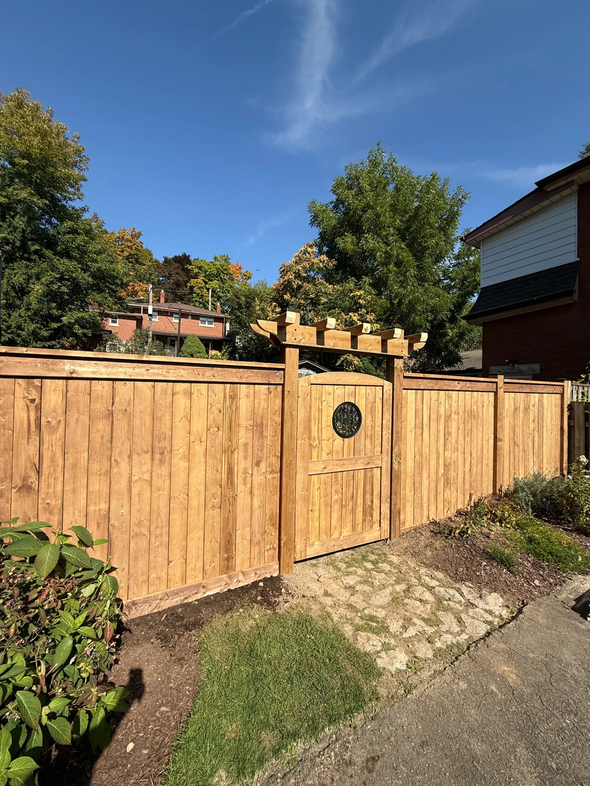Wooden fence with arbor gate, brown color. Clear blue sky, green trees in background.