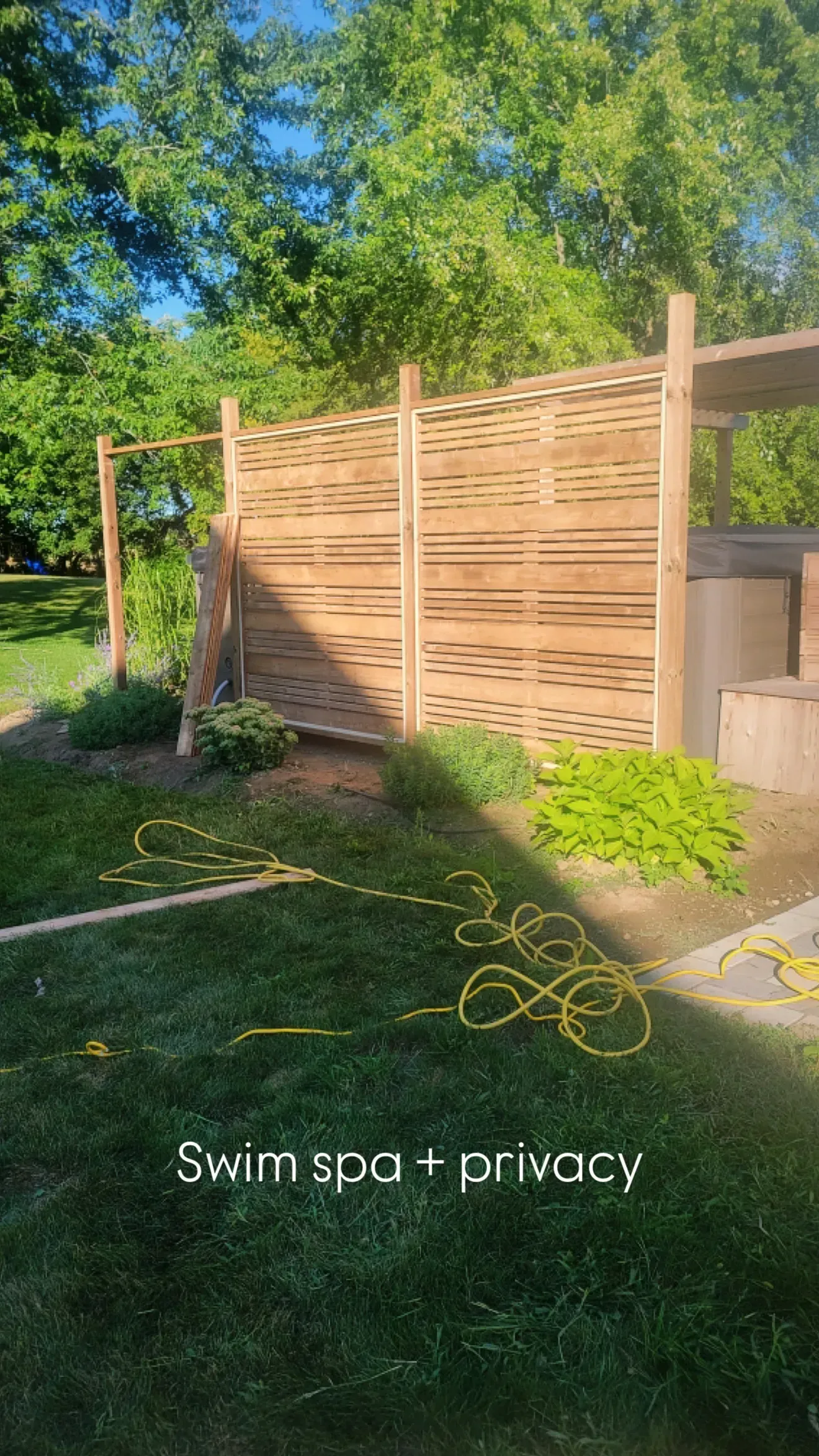 Wooden privacy screen encloses swim spa. Brown wood, green plants, and lawn in sunny outdoor setting.