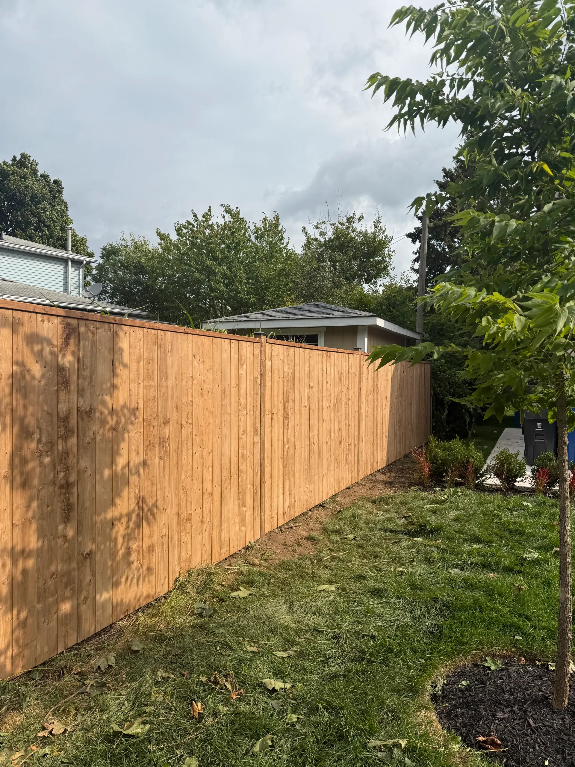 Wooden privacy fence along a green lawn with trees and a cloudy sky.