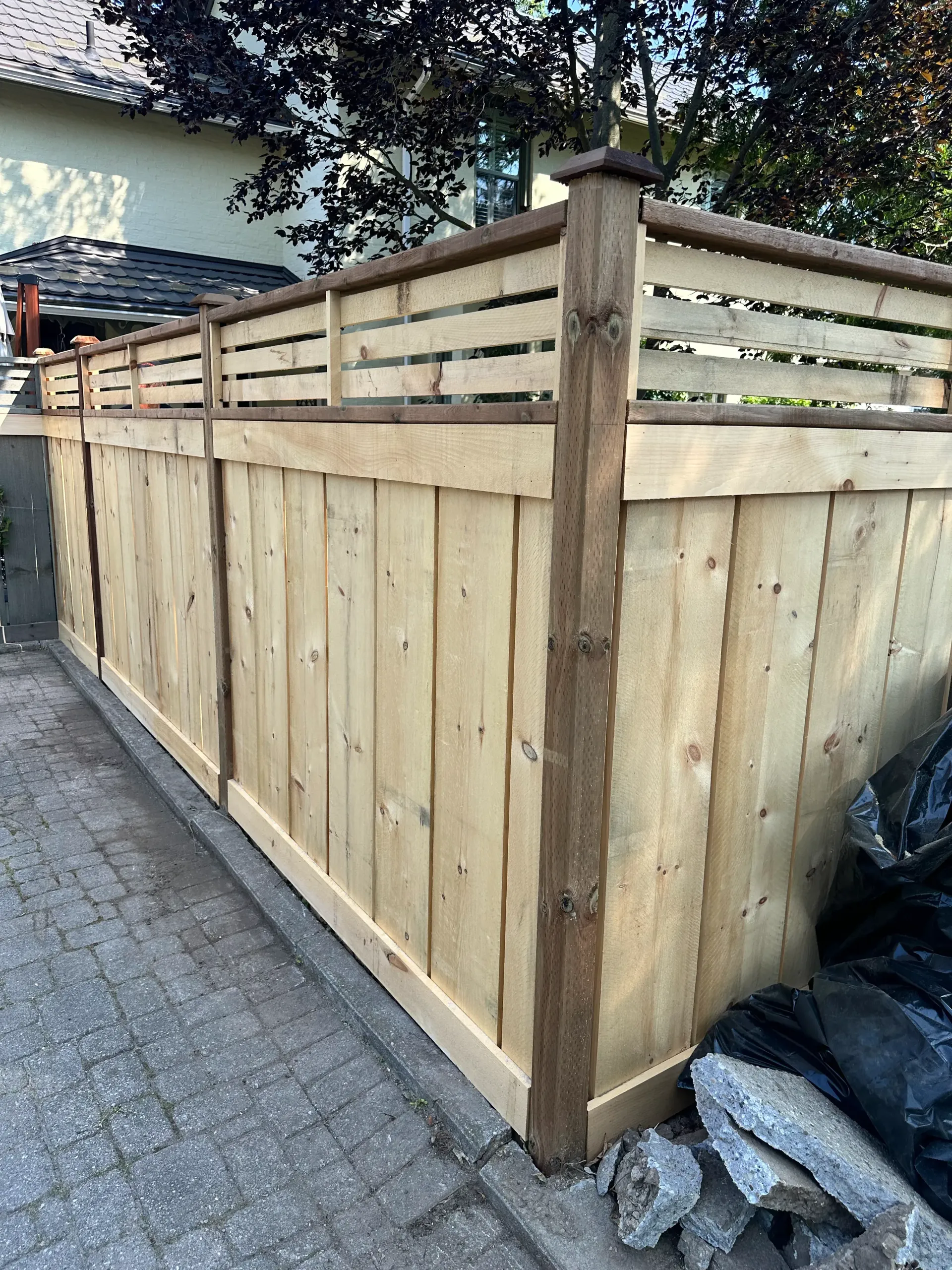 Wooden fence with vertical planks and horizontal top slats. Brown posts and frame. Gray paving stones.