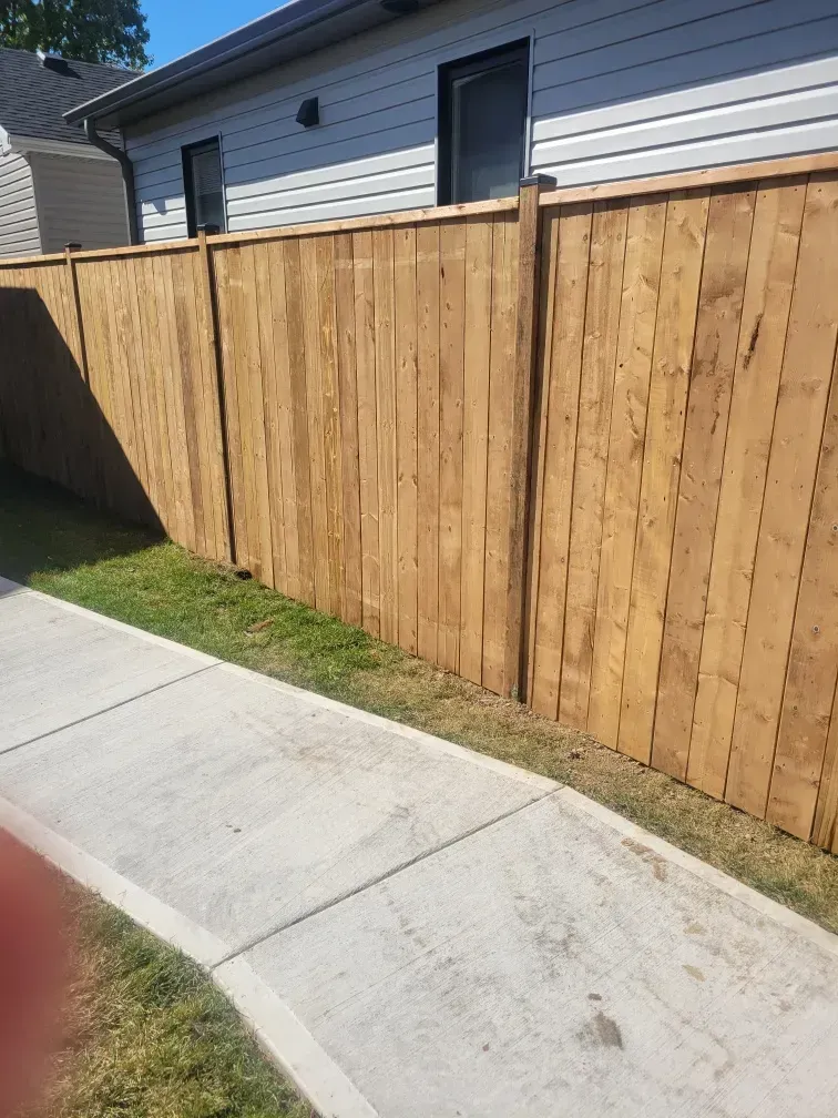 Wooden fence next to a sidewalk and grass, with a building in the background.