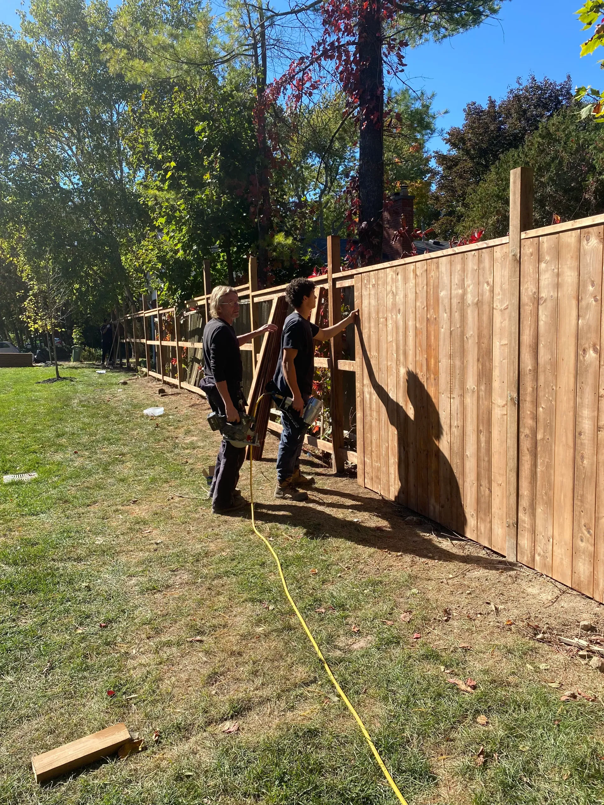 Two people building a wooden fence outdoors on a sunny day.