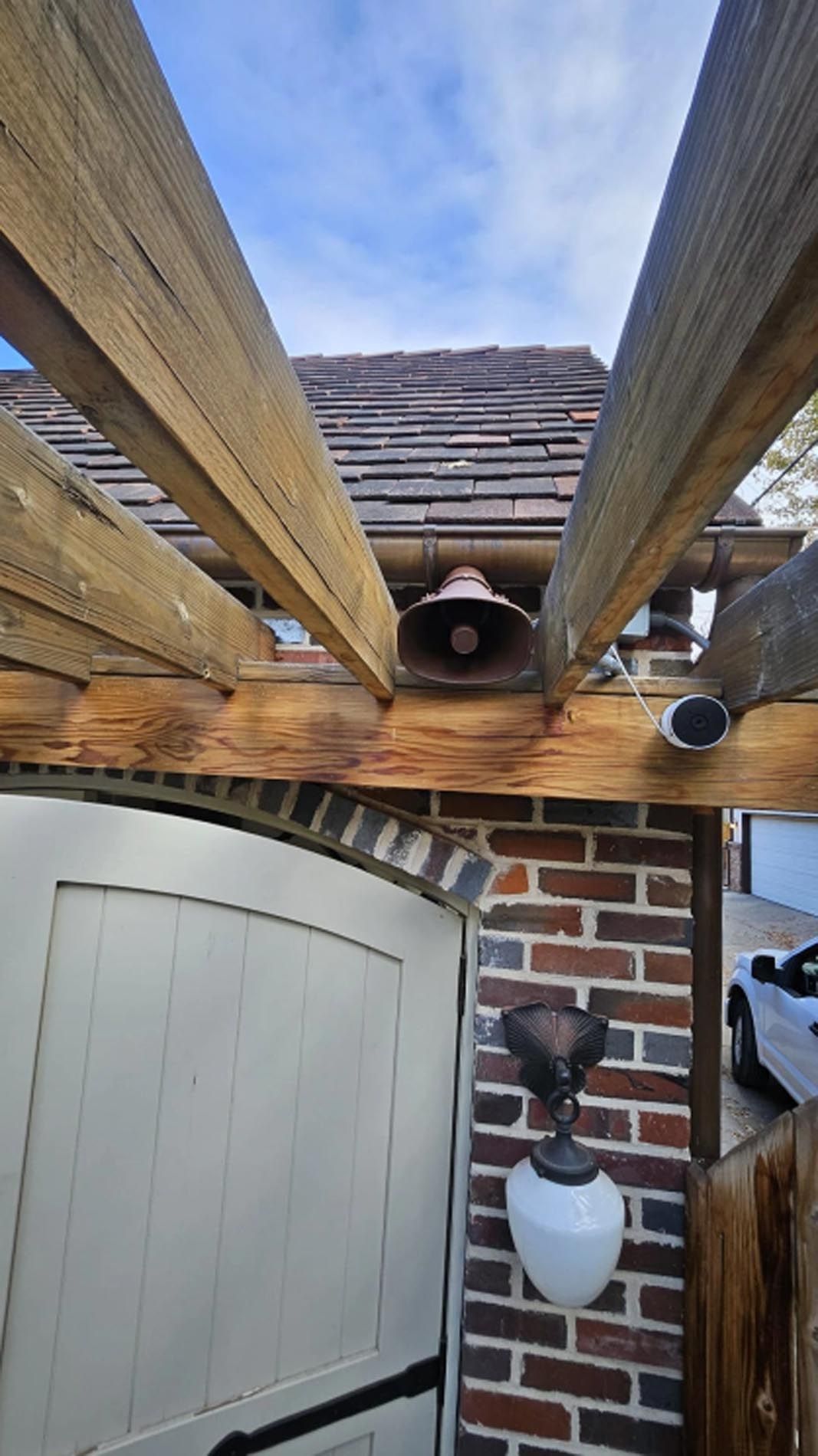 A white car is parked under a wooden pergola next to a brick wall.