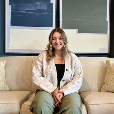 A smiling person with wavy hair sits on a beige couch wearing a cream cardigan and green pants, in front of abstract art.