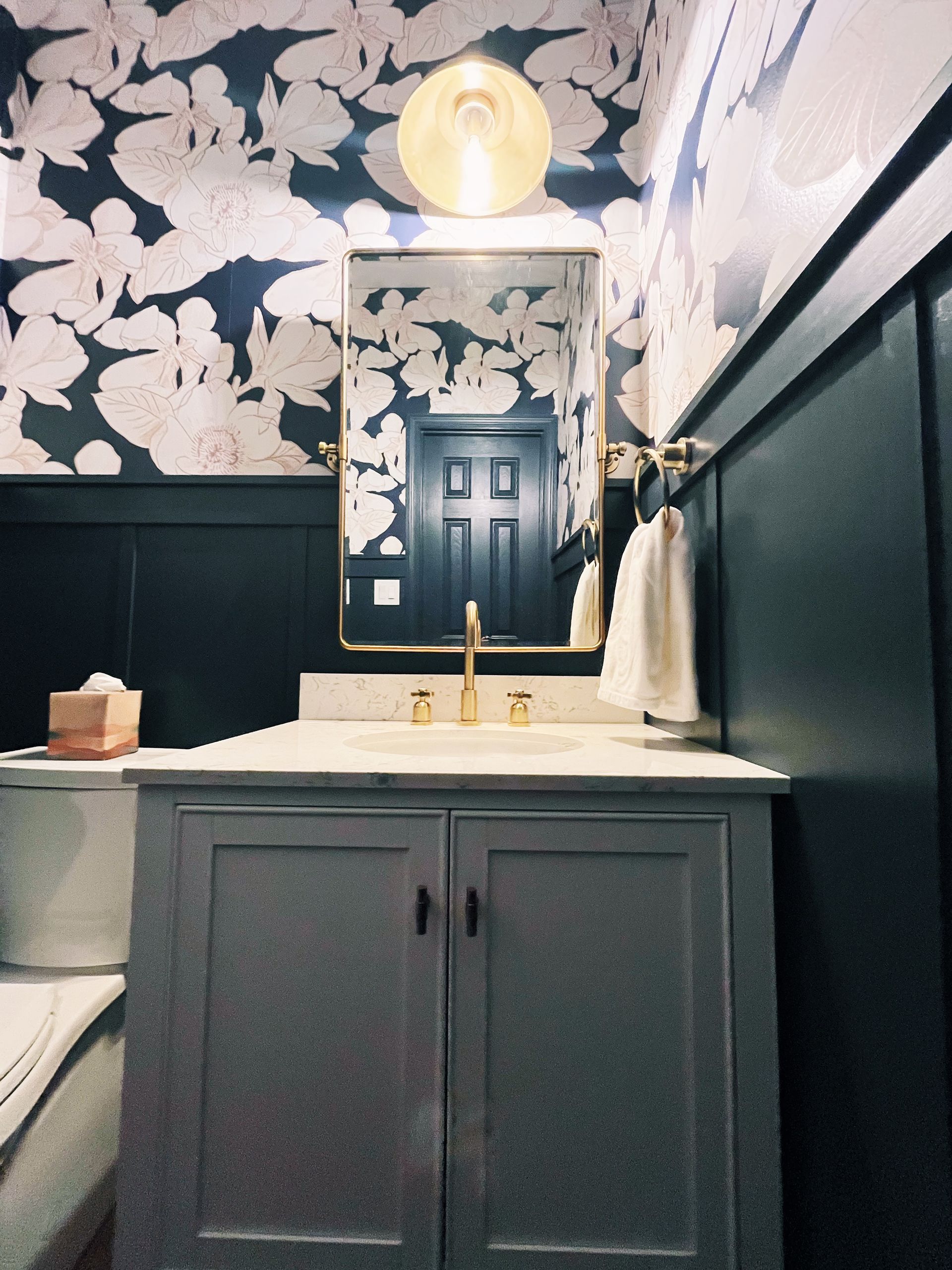 Bathroom vanity with marble countertop, mirror, and brass faucet against dark paneling and floral wallpaper.
