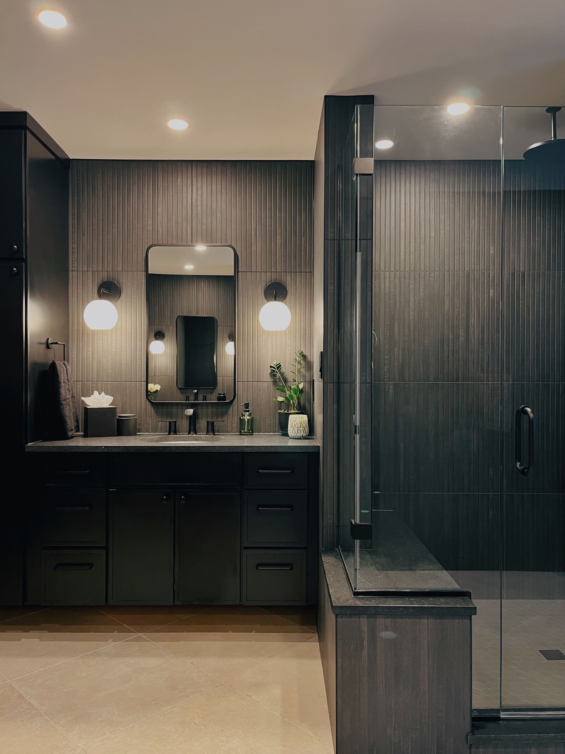 Modern dark bathroom with double vanity, round mirrors, wall sconces, and a glass shower.