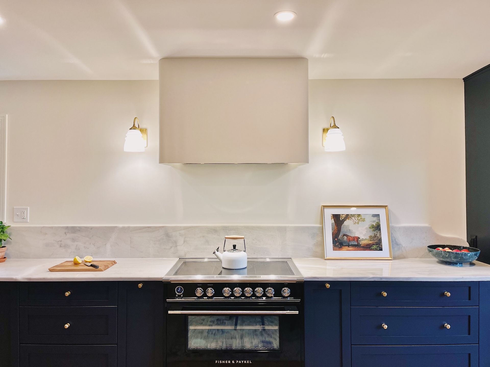 A modern kitchen featuring navy blue cabinets, a marble backsplash, a stainless steel oven, and a beige range hood.