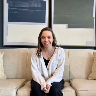A person sits smiling on a light-colored sofa in front of two large, abstract, multi-toned paintings.