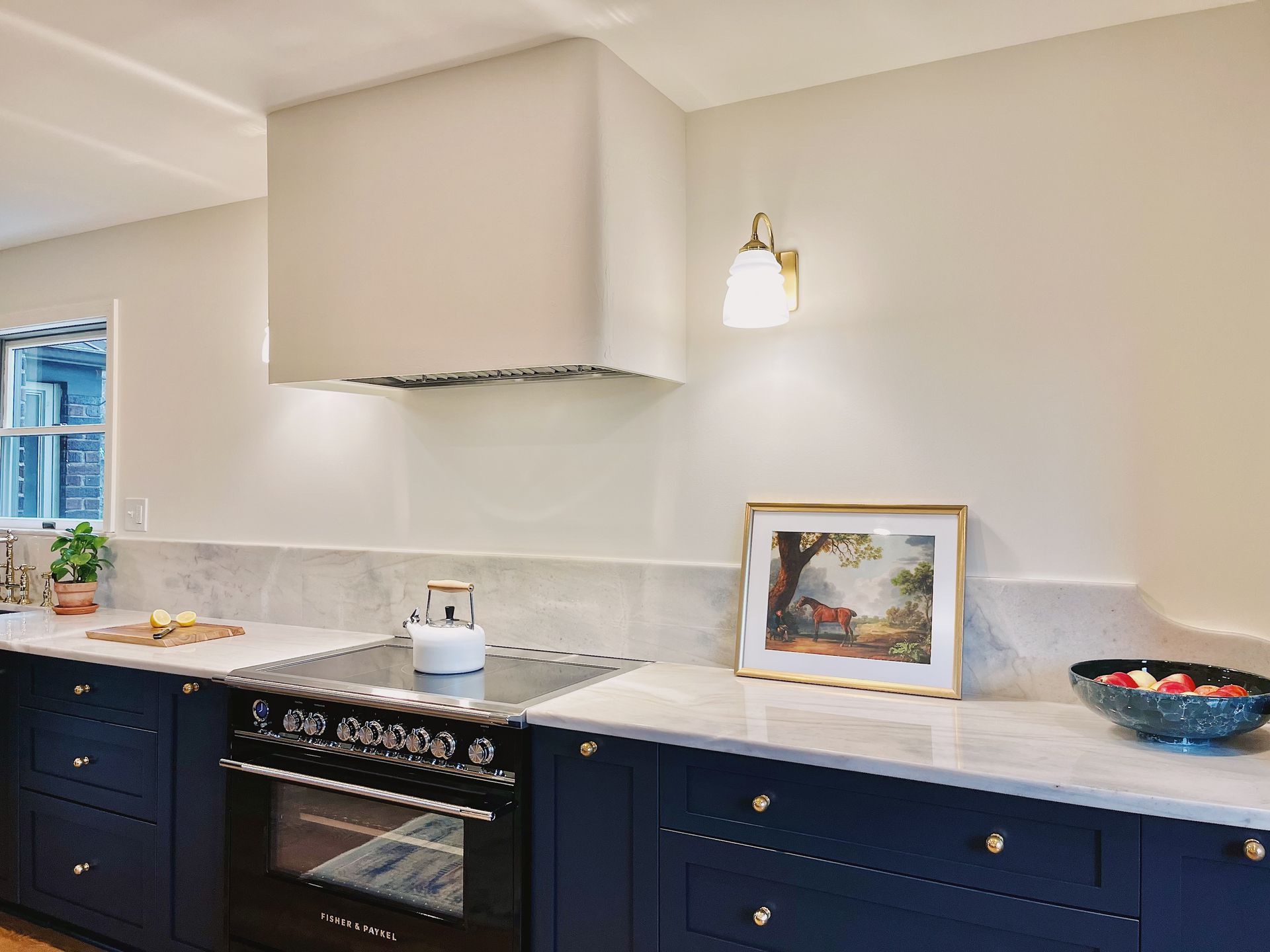 A kitchen with dark navy cabinets, a large white range hood, a marble countertop, and a framed landscape painting.