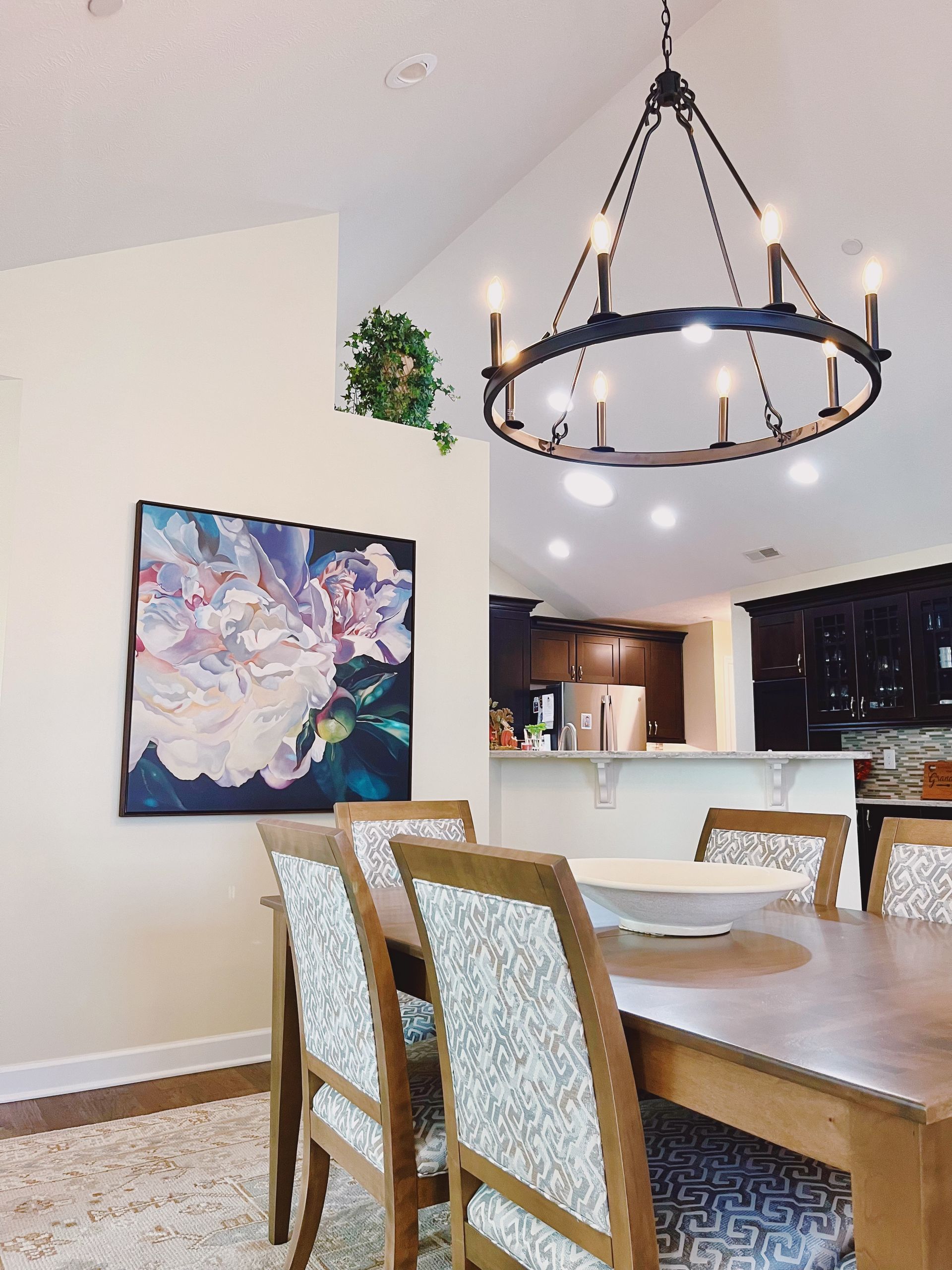 A dining room featuring a wooden table with patterned chairs under a large circular chandelier, with art on the wall.