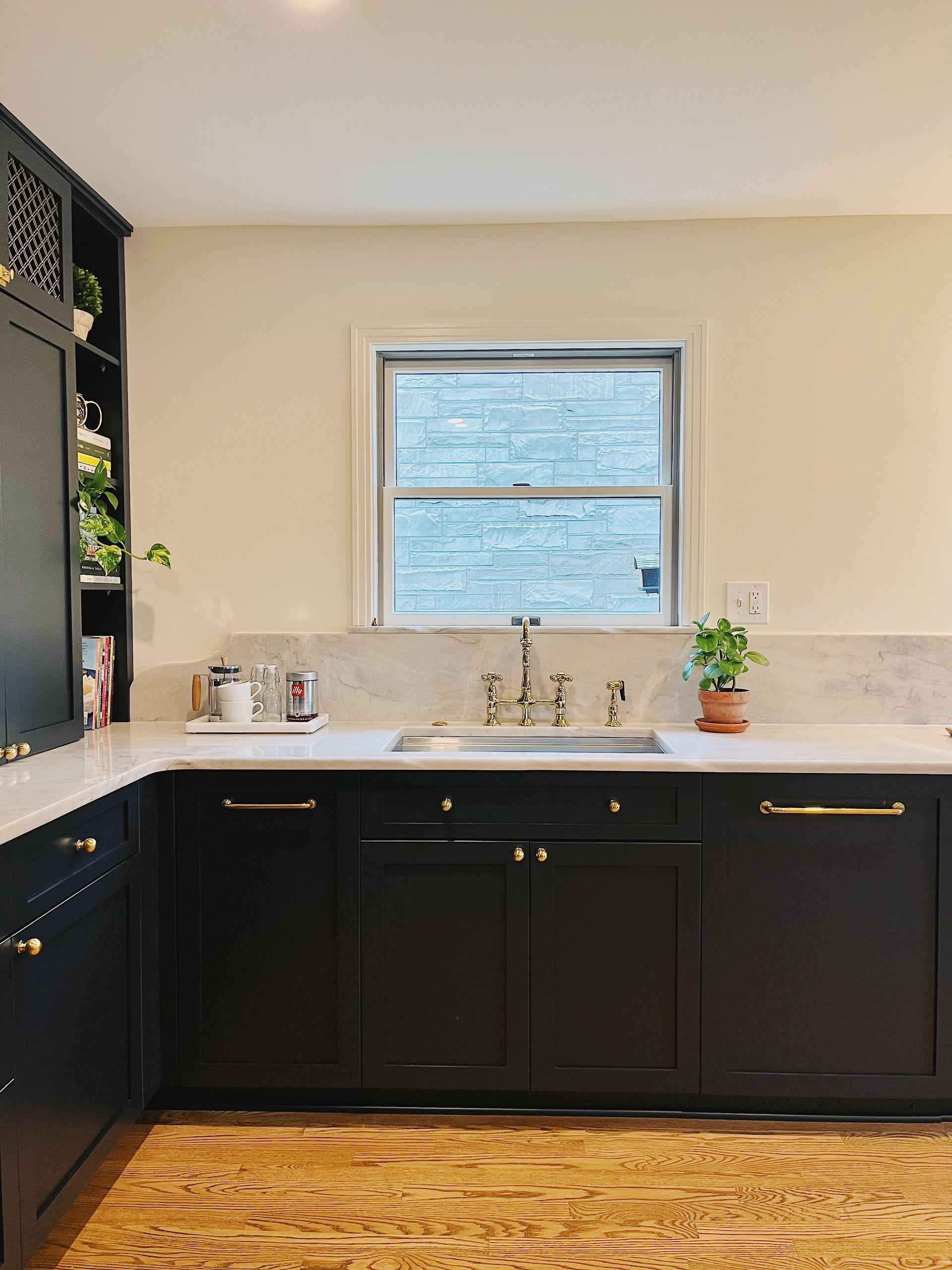 A kitchen scene featuring black cabinets, a marble backsplash, a sink under a window, and light-colored wooden flooring.