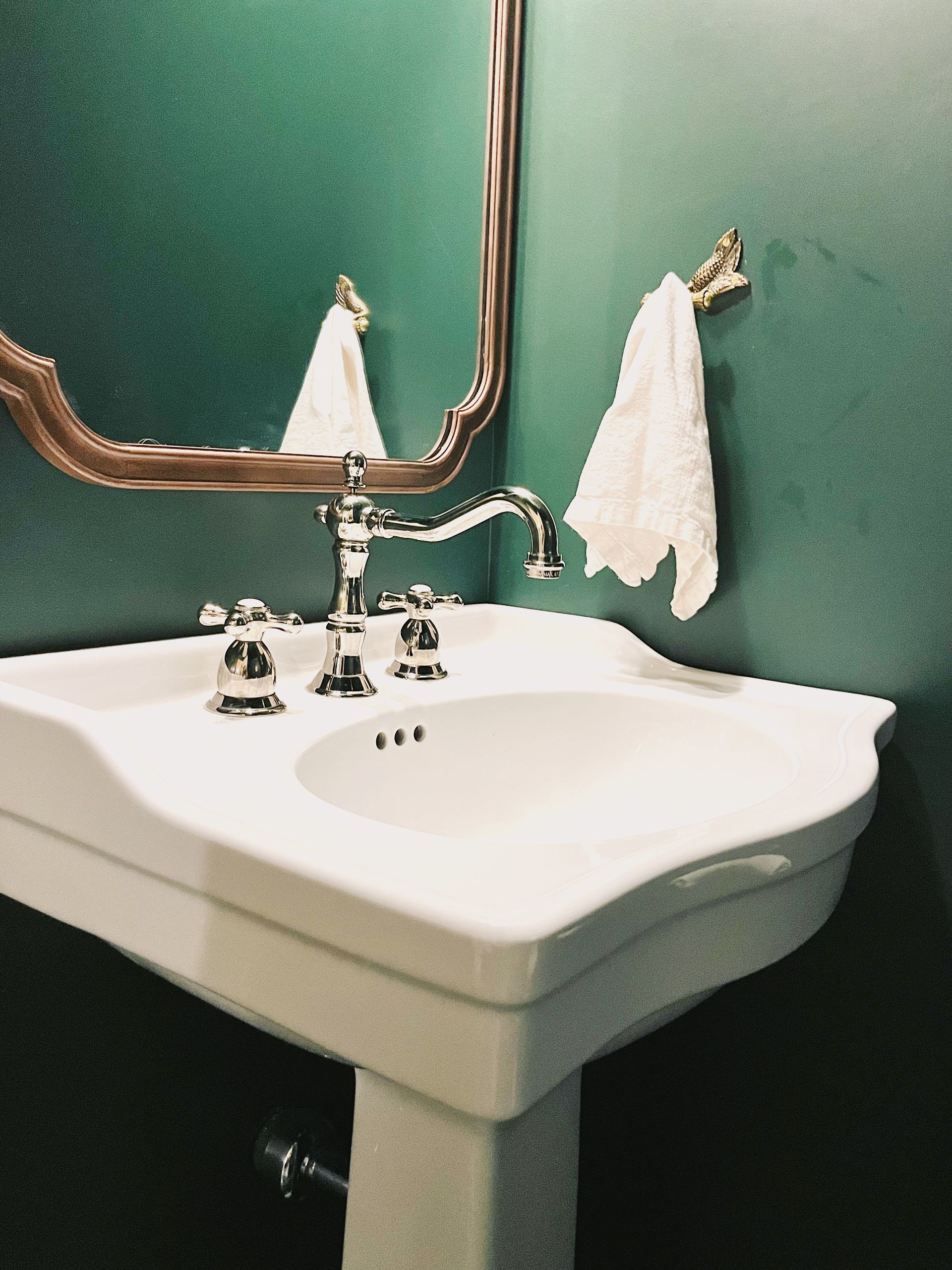 A white pedestal sink with silver fixtures stands against dark green walls below a decorative mirror with a gold frame.