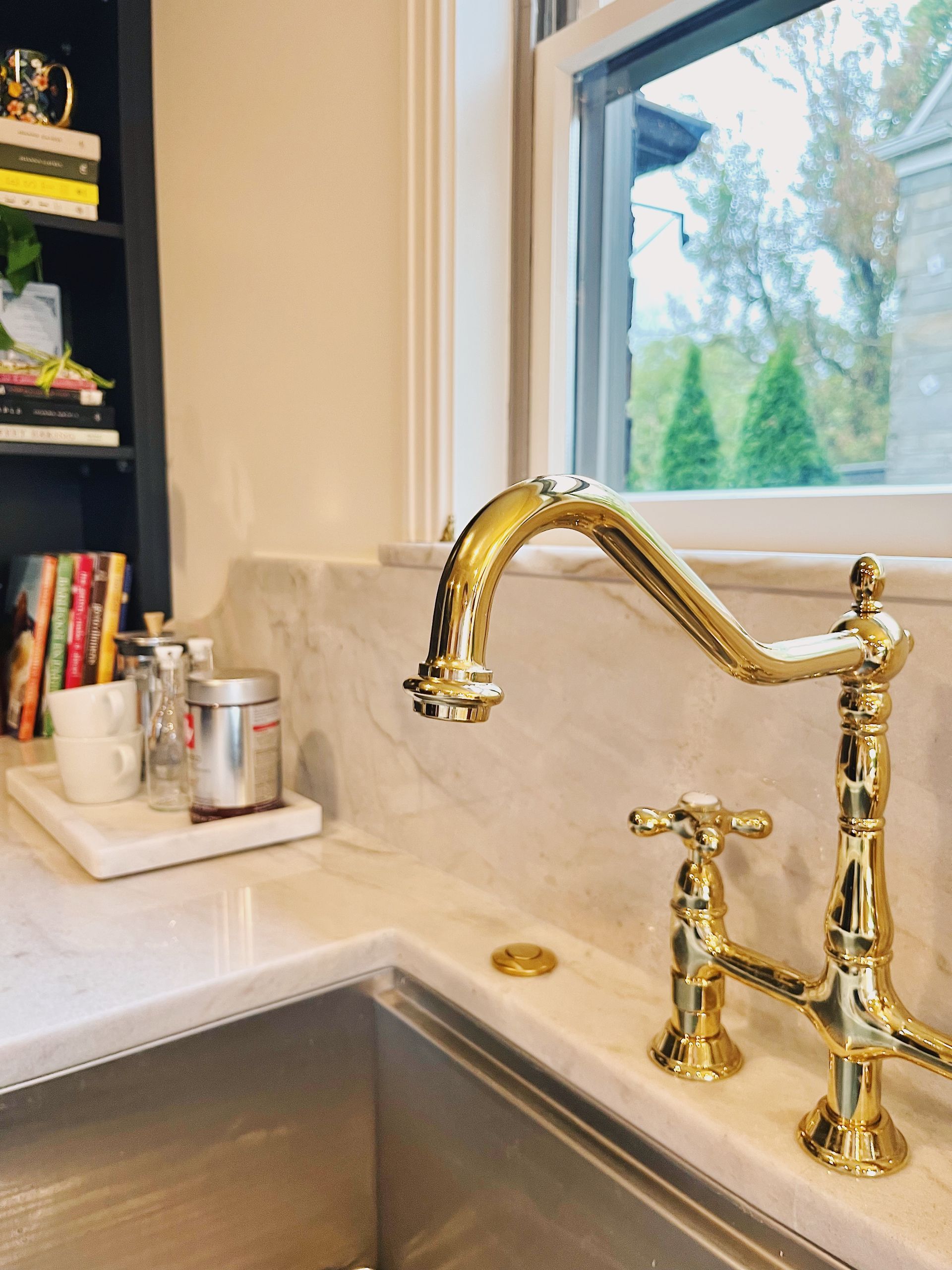 A polished gold bridge-style faucet stands on a white marble countertop by a window, with a partial shelf of books nearby.