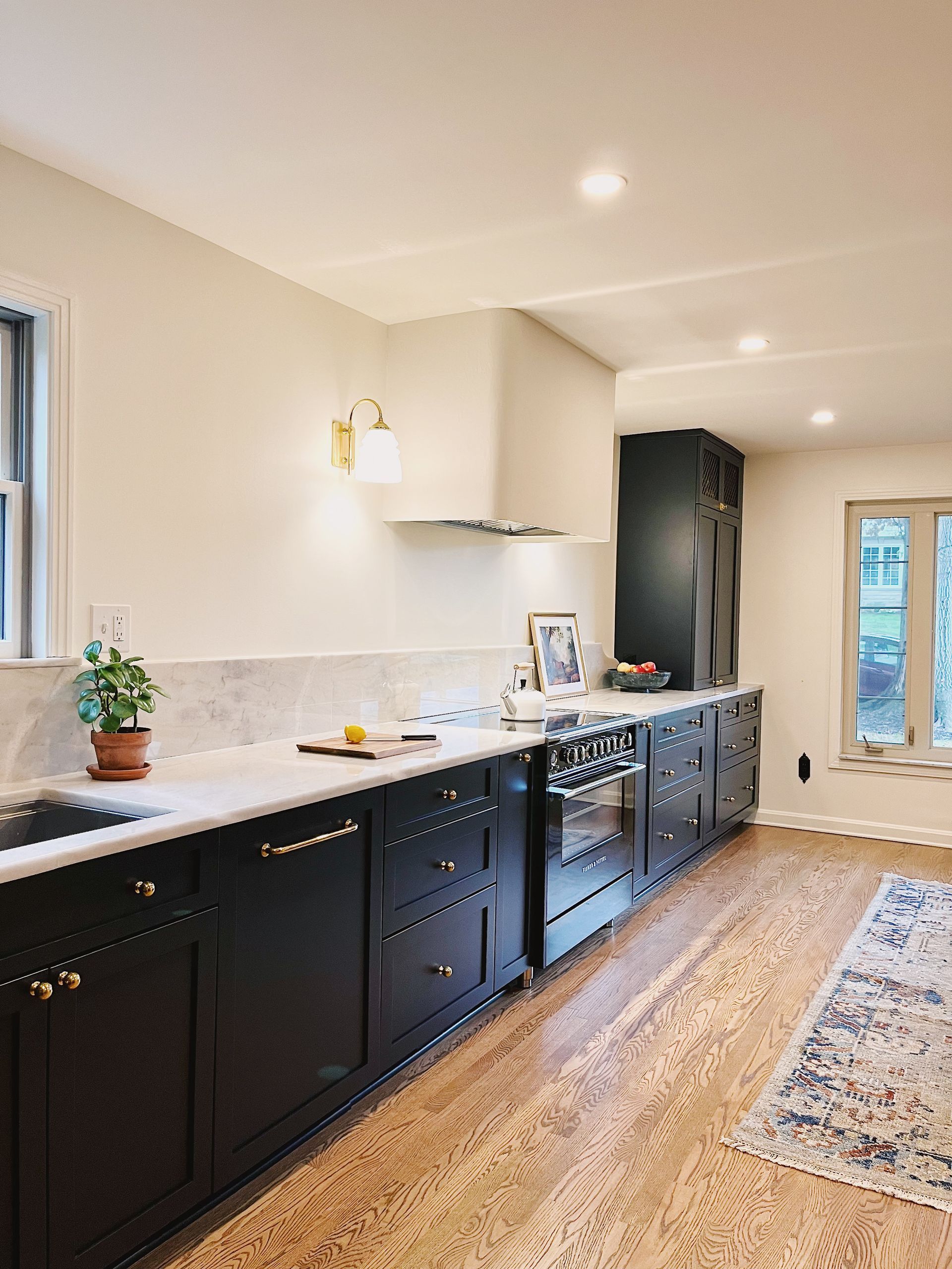 A modern kitchen featuring black cabinetry, white marble countertops, a custom vent hood, and natural wood flooring.