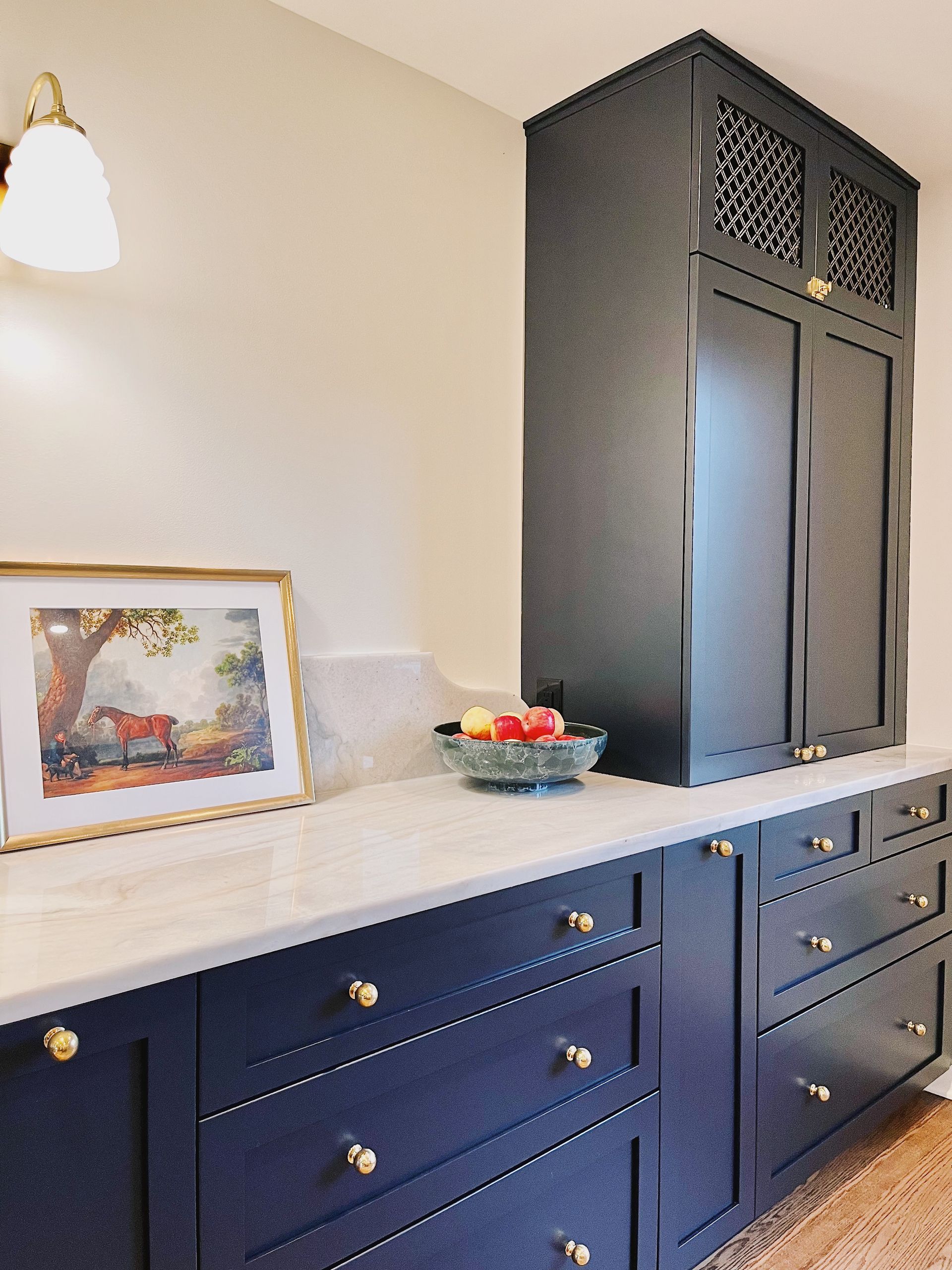 Dark blue cabinetry with gold hardware, a white marble countertop, a bowl of fruit, and a framed painting on a wall.