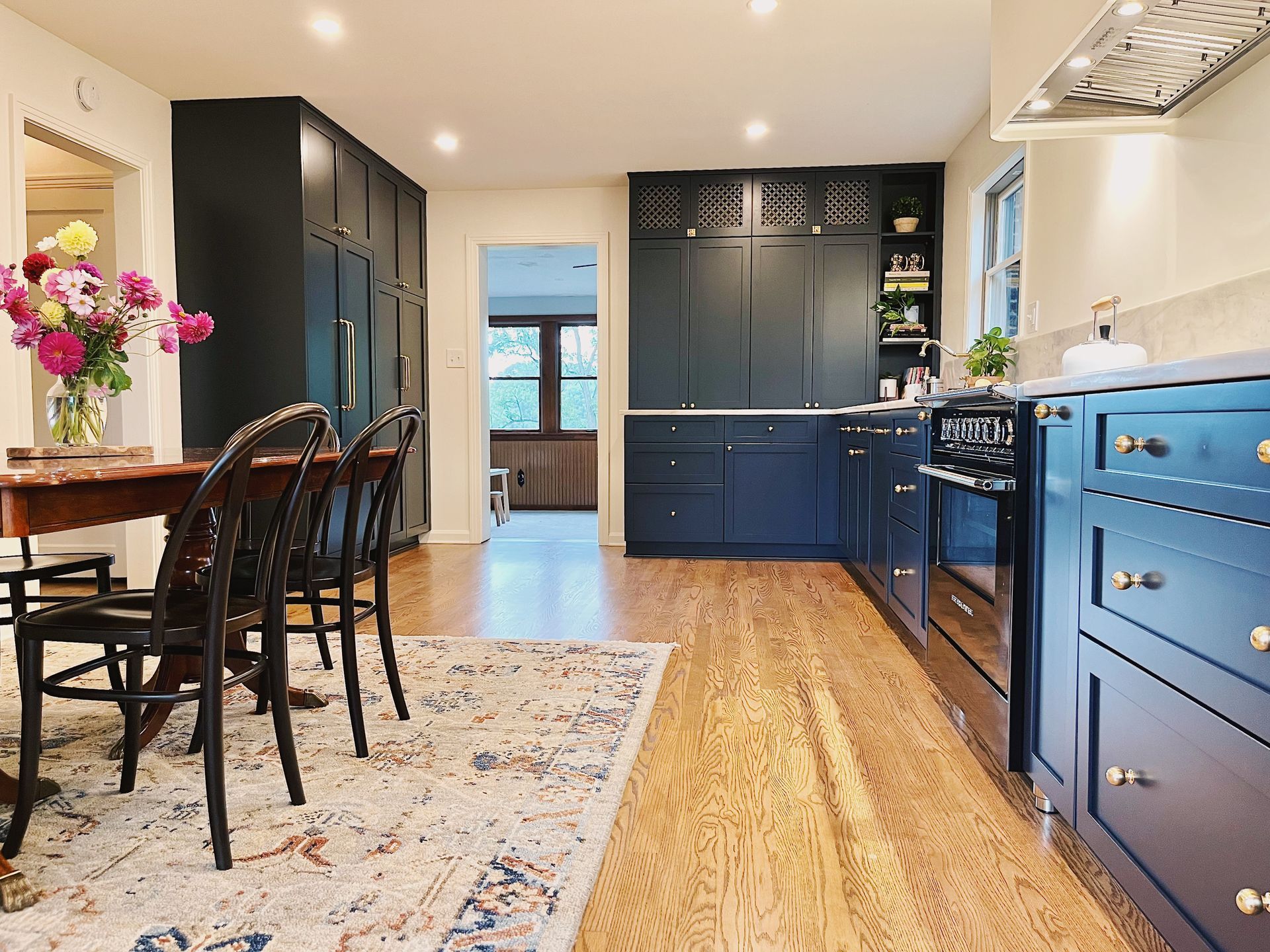 A kitchen featuring dark blue cabinets, hardwood floors, a patterned area rug, and a dining table with black chairs.