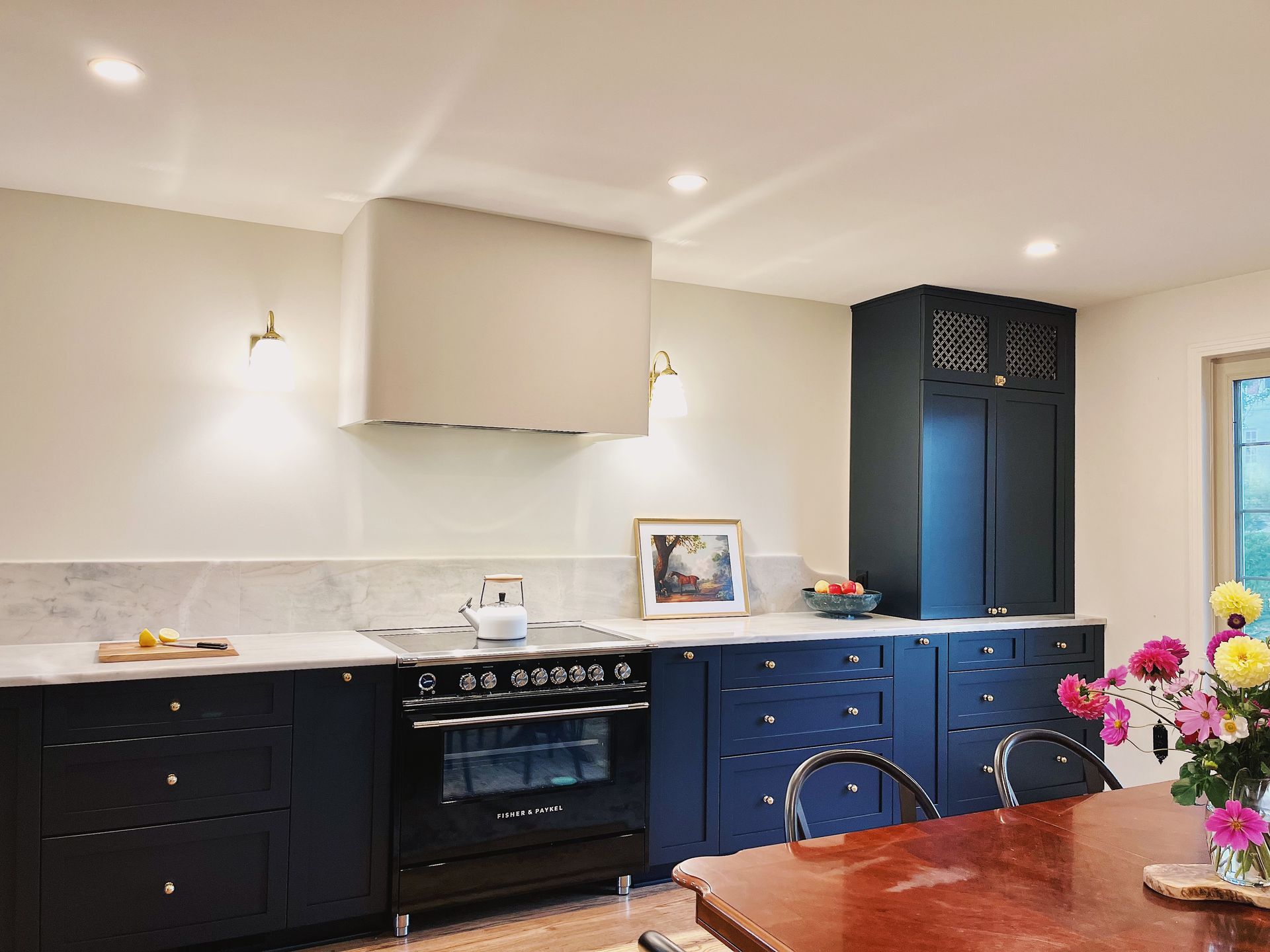 A dark blue kitchen with marble countertops, a central stove, a large range hood, and a wooden dining table with flowers.
