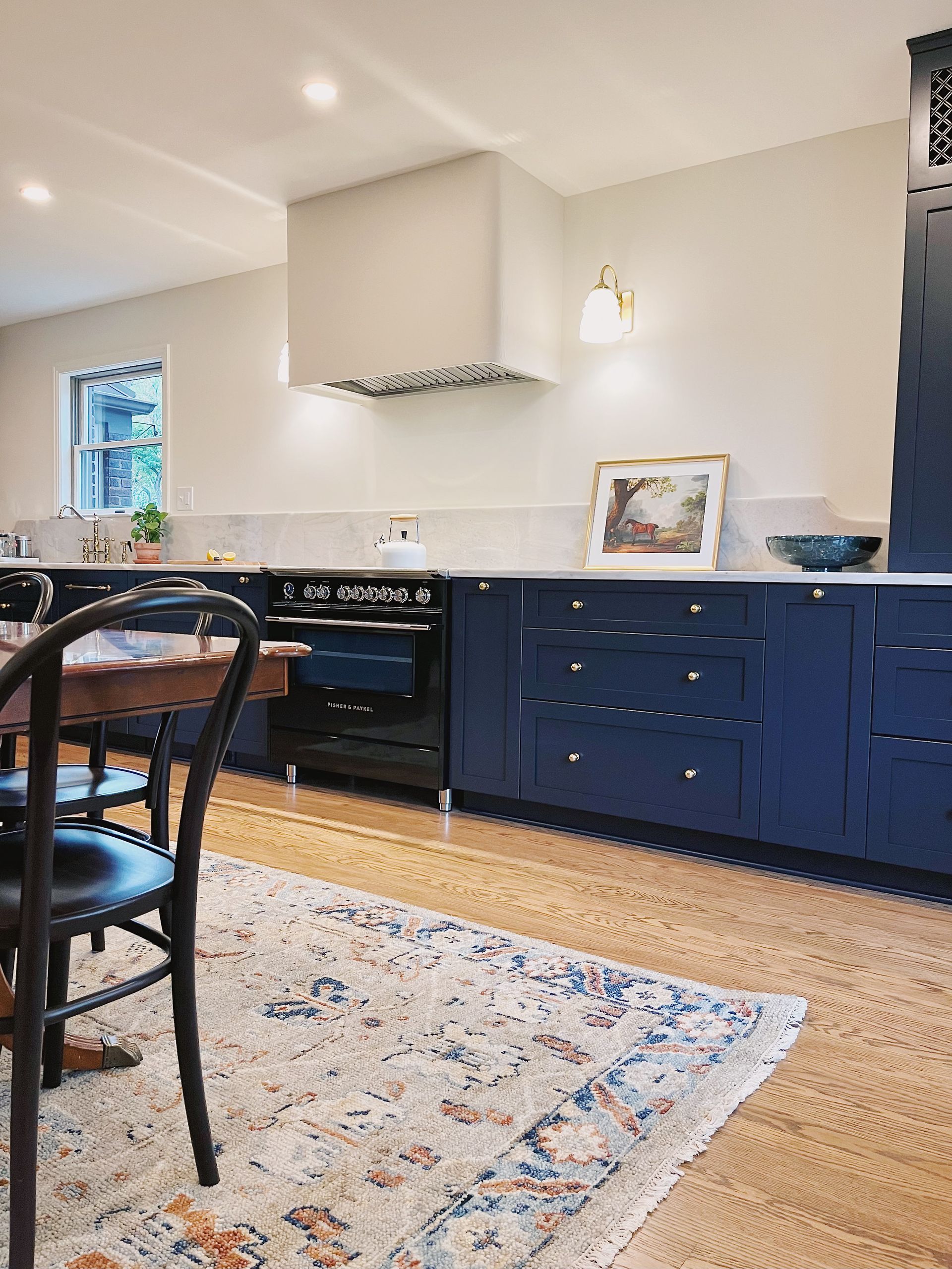 A kitchen with dark navy cabinets, a white range hood, a black stove, and a patterned area rug on light hardwood floors.
