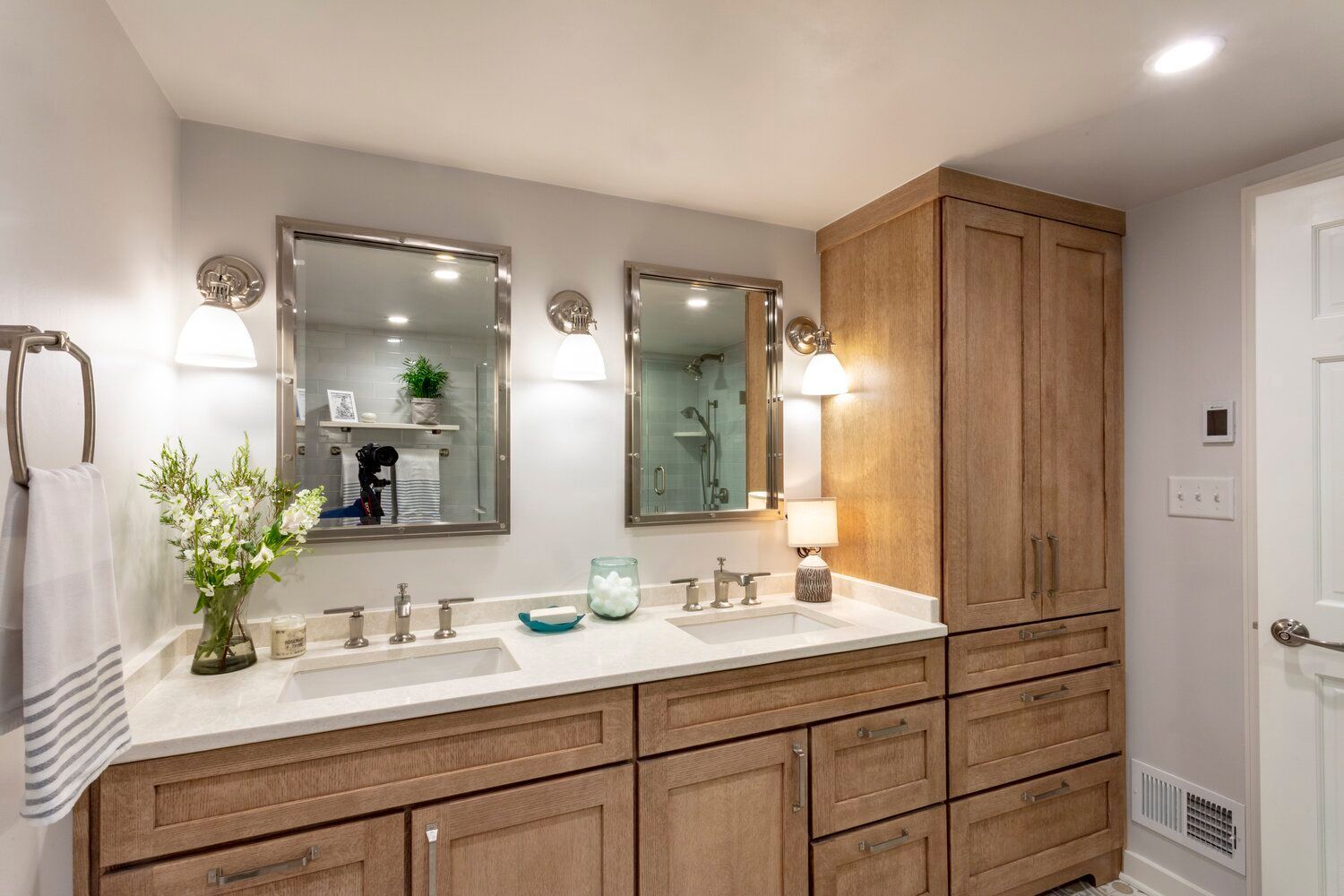 A double-sink bathroom vanity with light wood cabinetry, white countertops, two framed mirrors, and wall sconces.