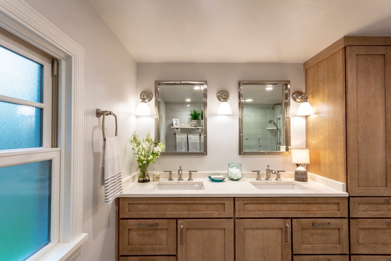 A bright bathroom vanity with wood cabinets, two sinks, two mirrors, and wall-mounted lights, next to a window.