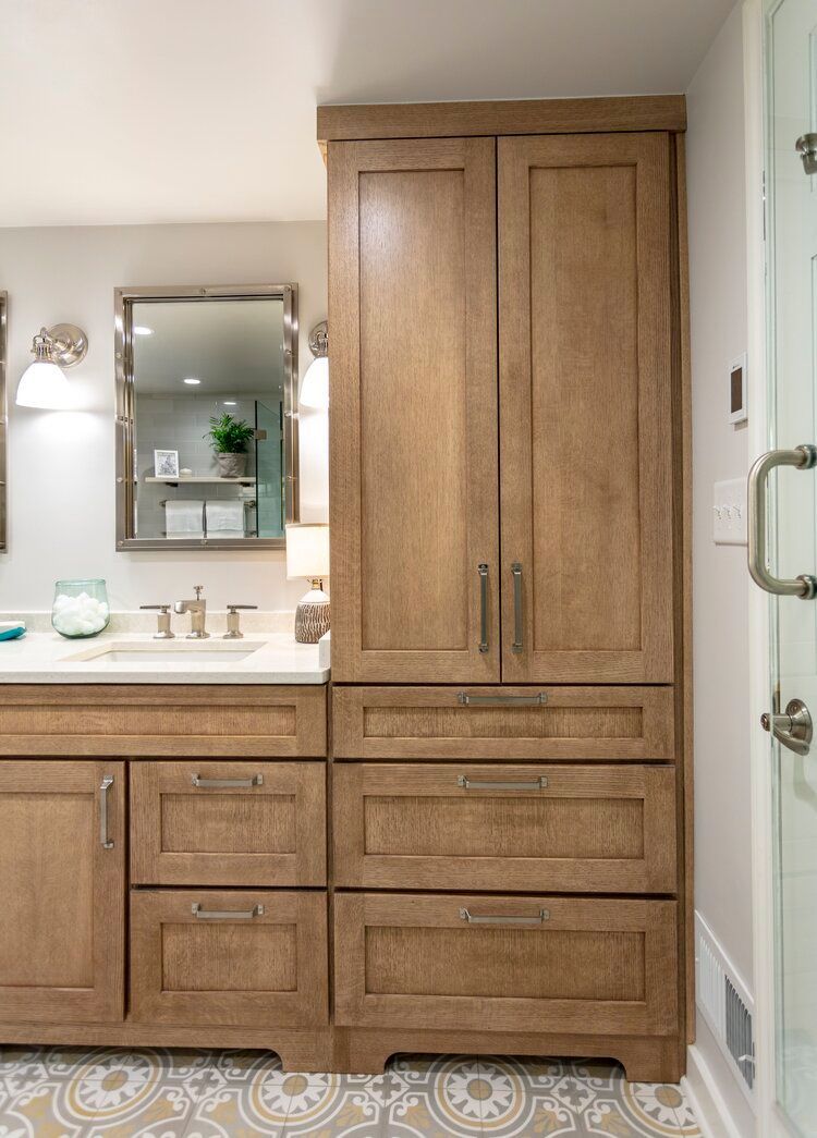 Light-toned wooden bathroom vanity with a tall storage cabinet, drawers, and an oval mirror, set on patterned tile floors.