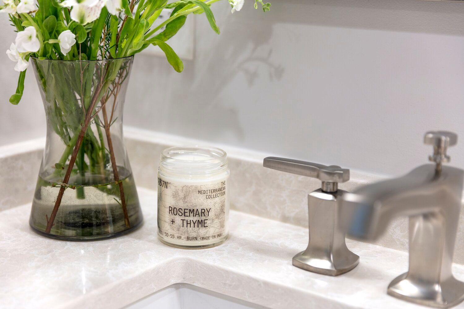 A glass vase with white flowers and a Rosemary & Thyme candle sit on a light marble bathroom vanity by a metal faucet.