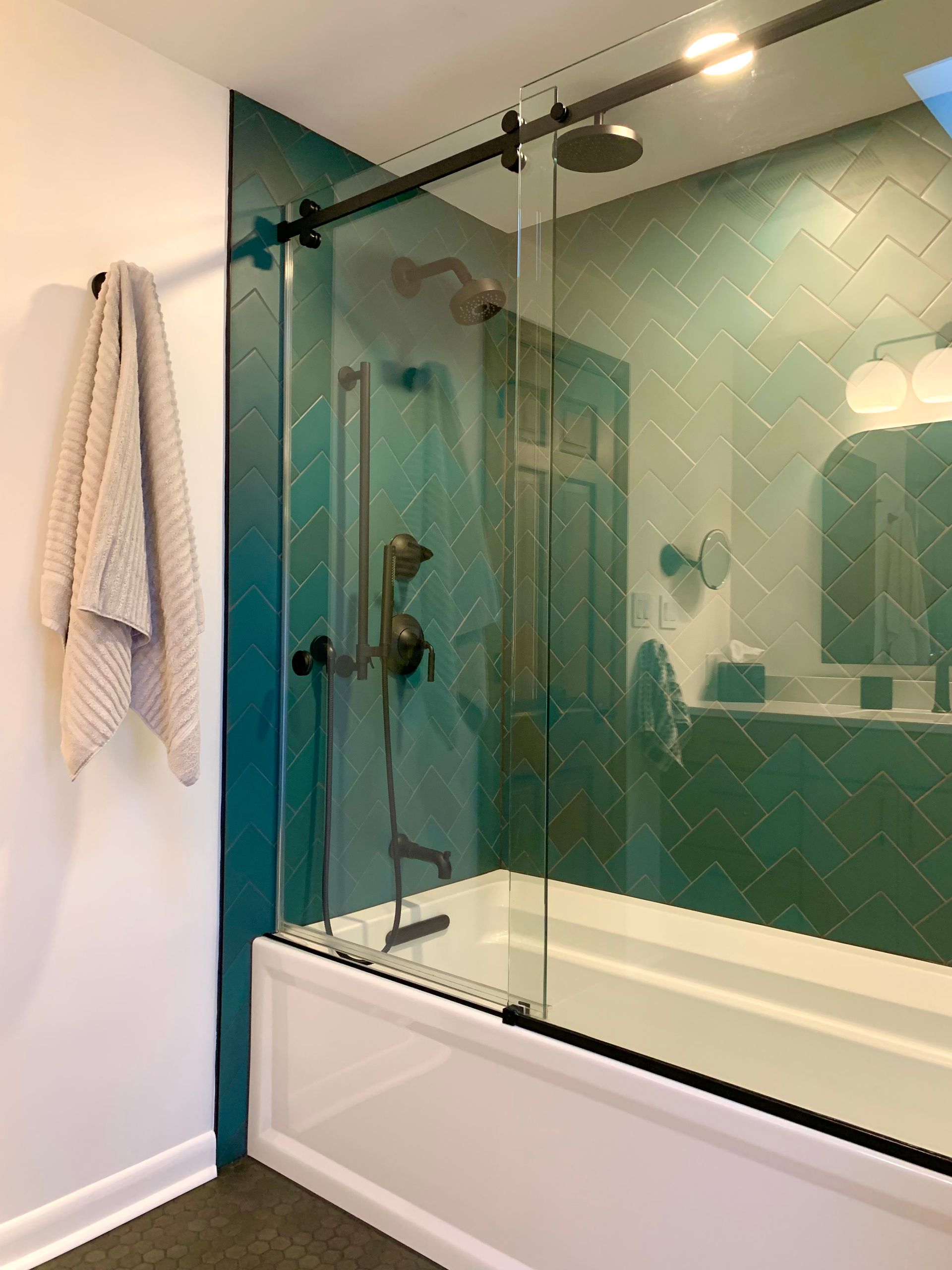 Glass shower enclosure with white tub, teal tile wall, and towel hanging on the left