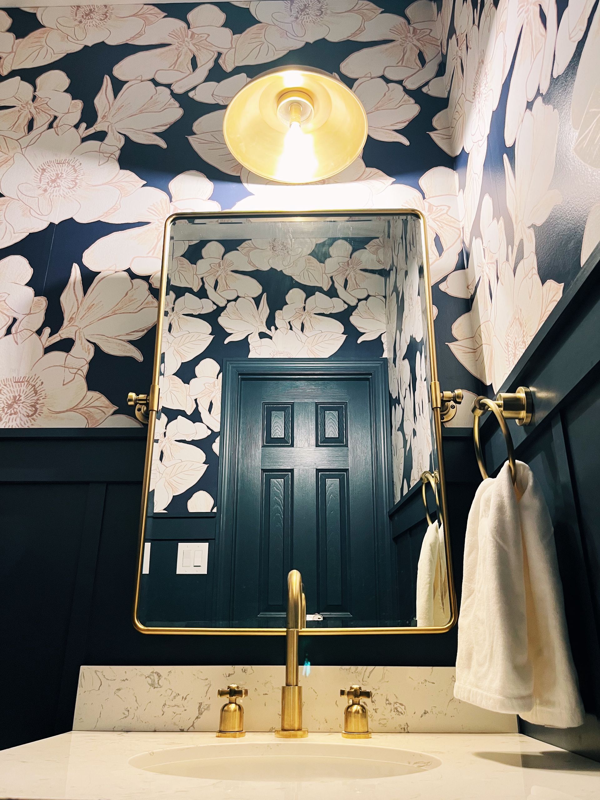 A powder room with navy floral wallpaper above dark wainscoting, a gold-rimmed mirror, gold faucet, and white countertop.
