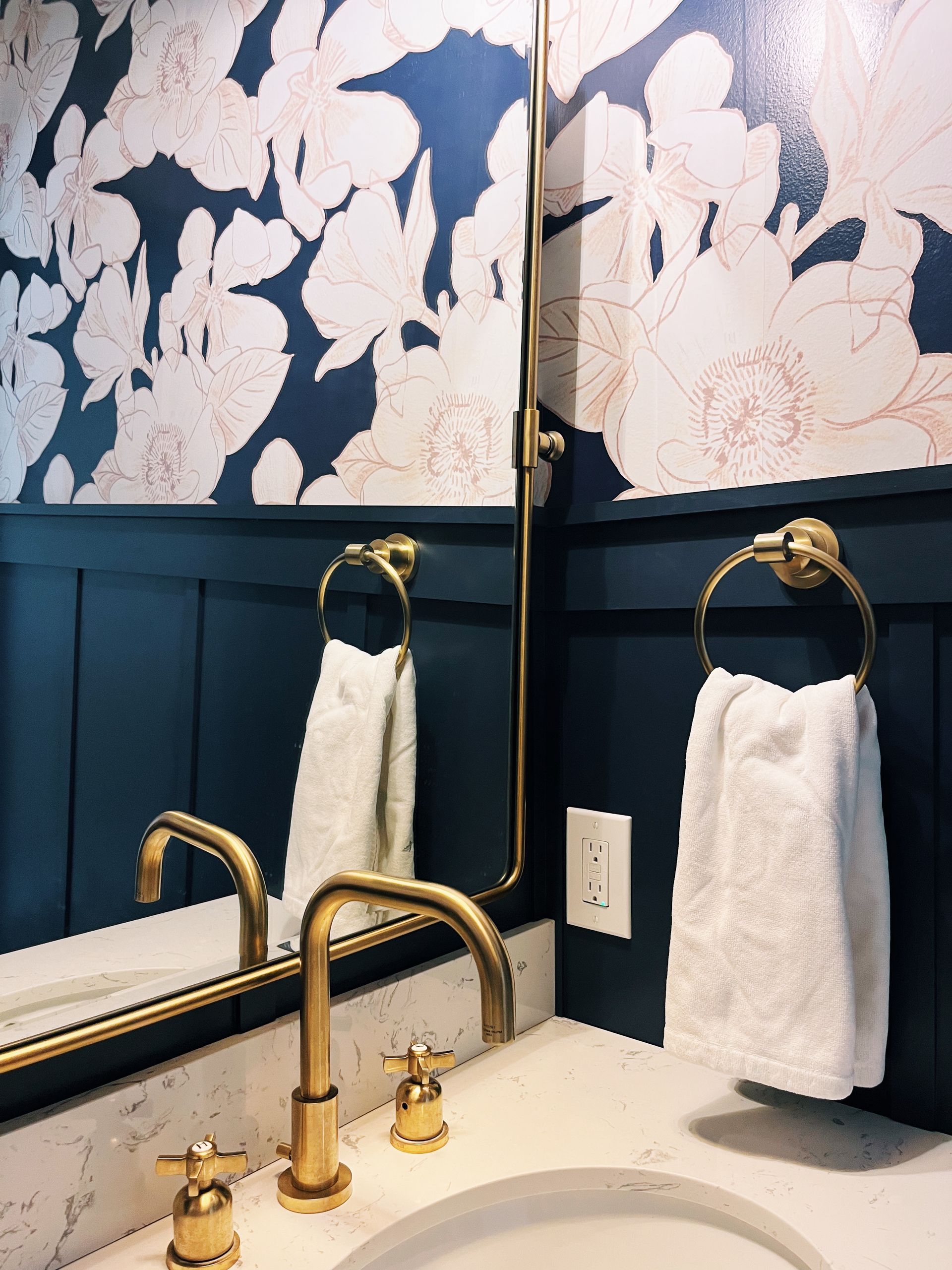 A bathroom vanity with navy paneled walls, floral wallpaper, brass faucet, and a white towel hanging on a brass ring.