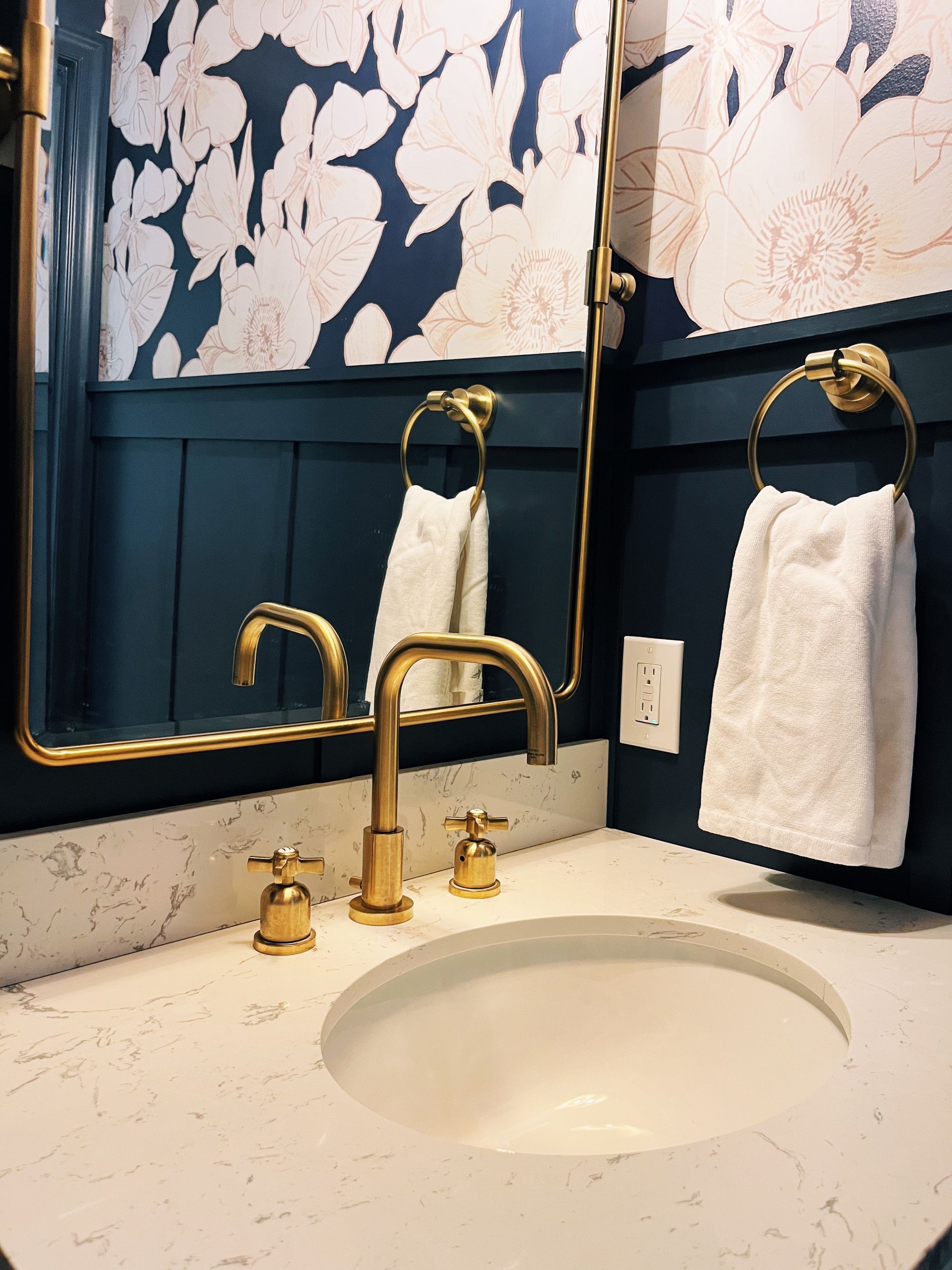 Bathroom vanity with gold faucet, white stone countertop, navy blue wainscoting, and floral wallpaper reflecting in mirror.