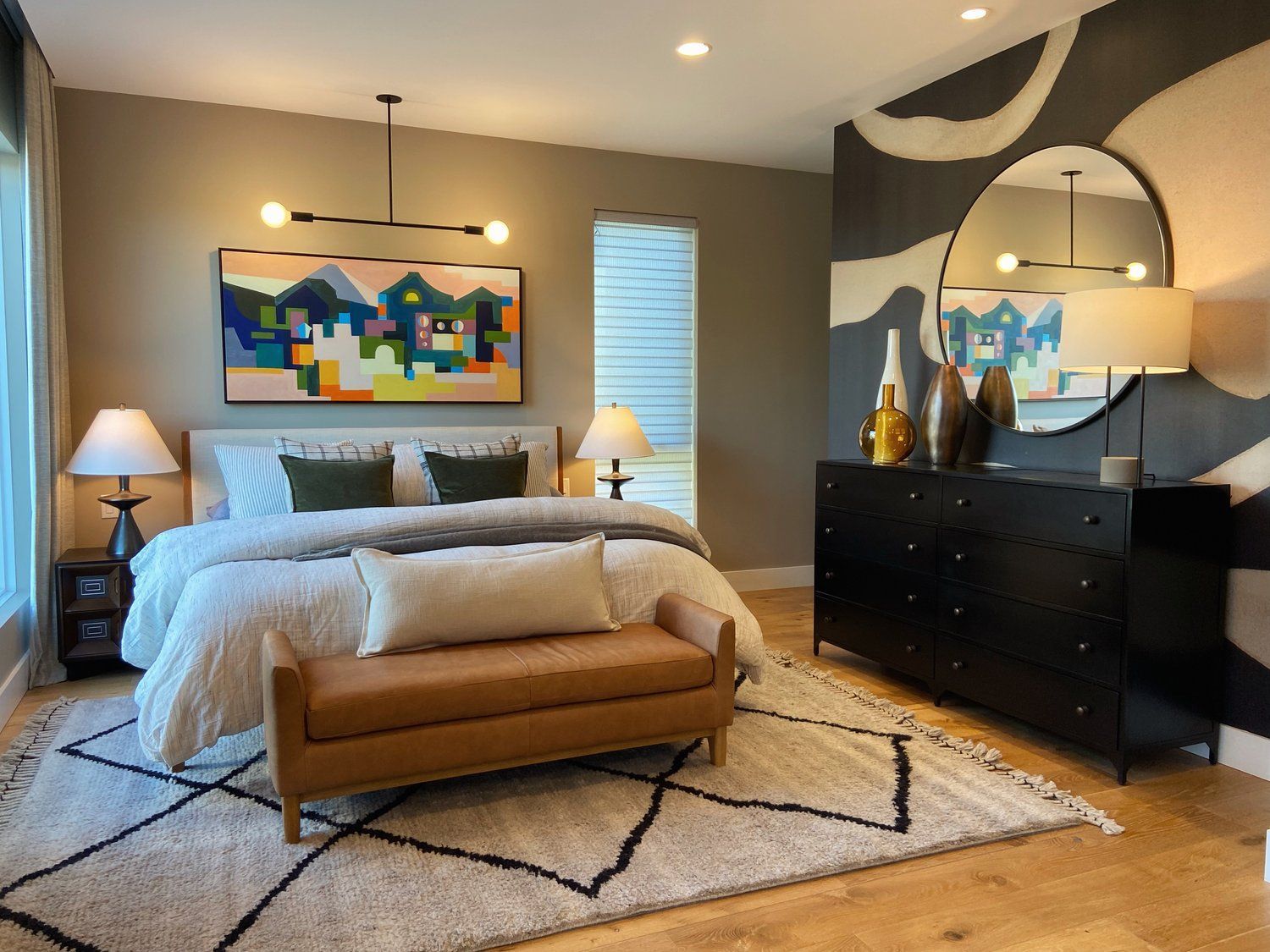 Modern bedroom with beige bed, bench, patterned rug, two lamps, dresser, and colorful wall art.