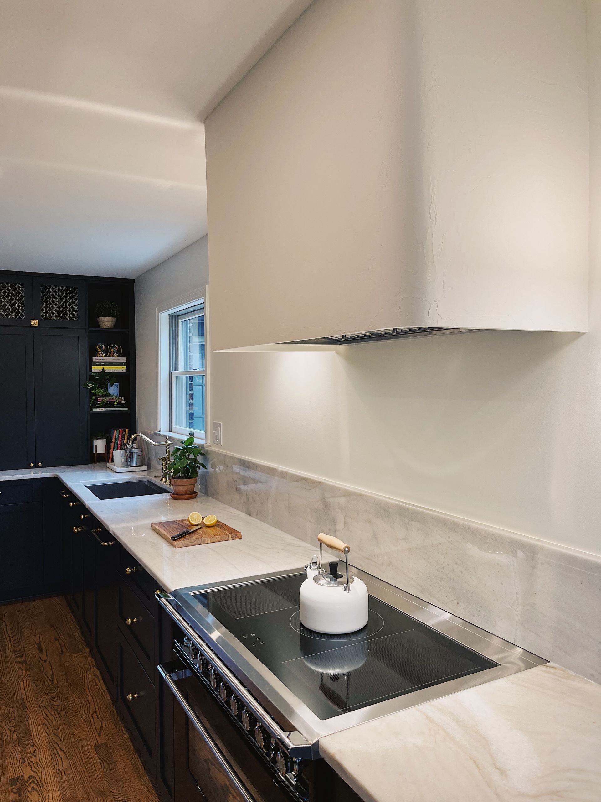 A sleek white stove hood hangs above a black range with a white kettle, light stone counters, and dark kitchen cabinets.