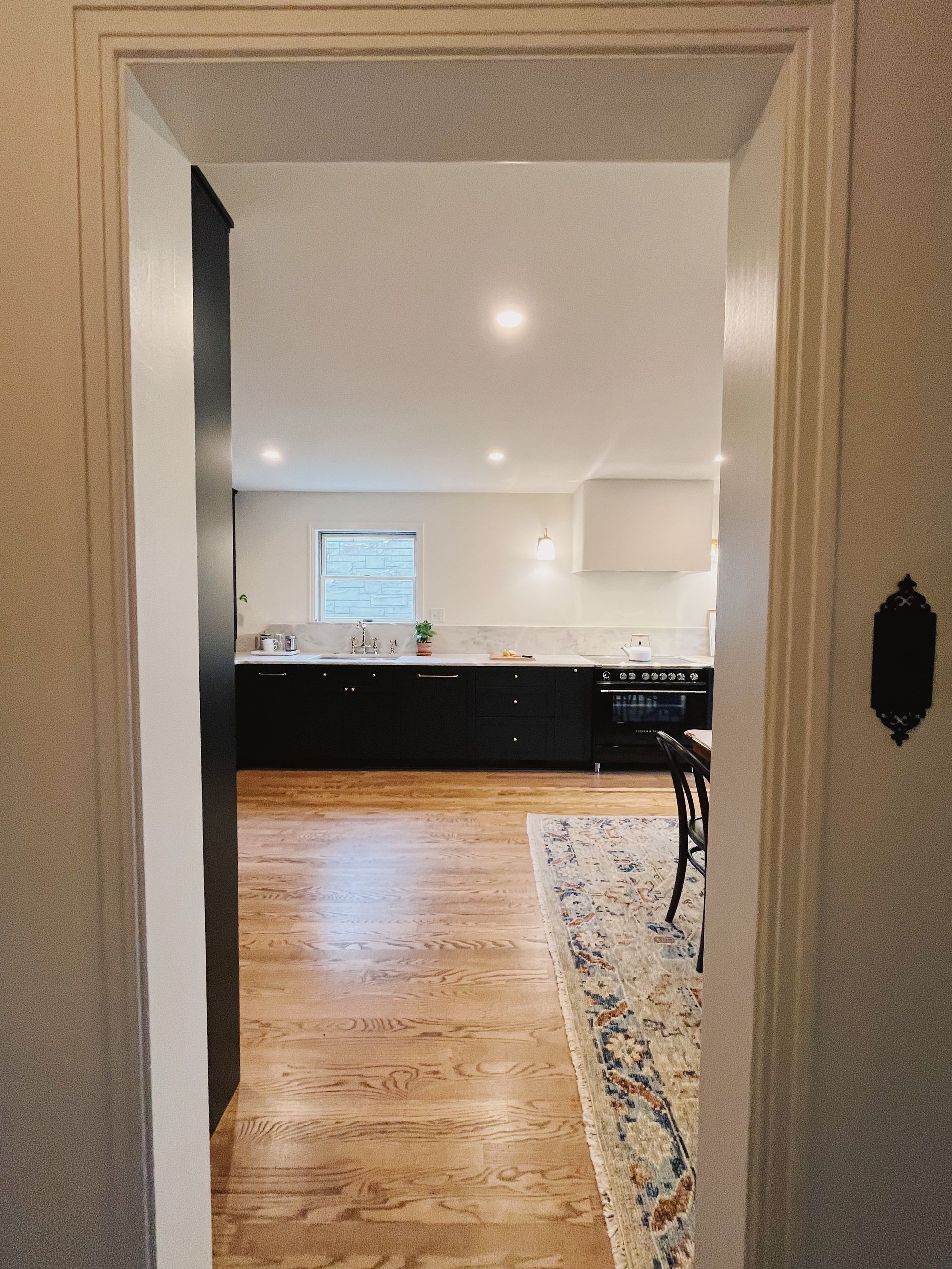 View through a doorway into a bright kitchen with dark cabinets, wood floors, and a patterned rug.