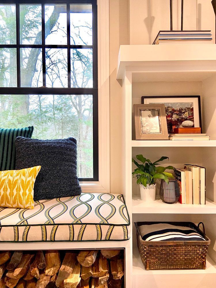 A window seat with colorful cushions, wood storage underneath, and white shelving displaying decor, books, and a basket.