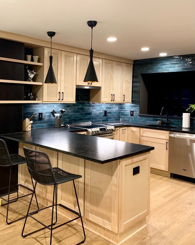 Modern kitchen with light wood cabinets, black countertops, blue backsplash tile, and two black pendant lights over a bar.