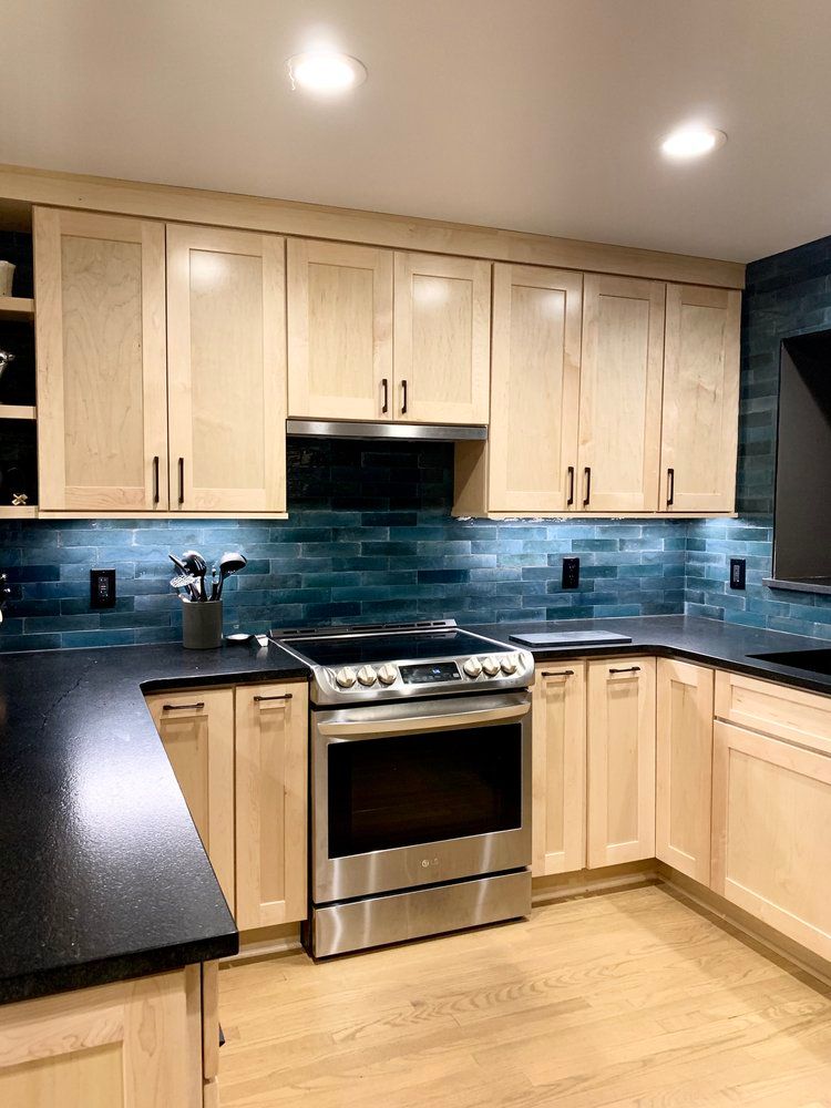 A kitchen with light-wood cabinets, black countertops, and a dark blue, rectangular-tiled backsplash.