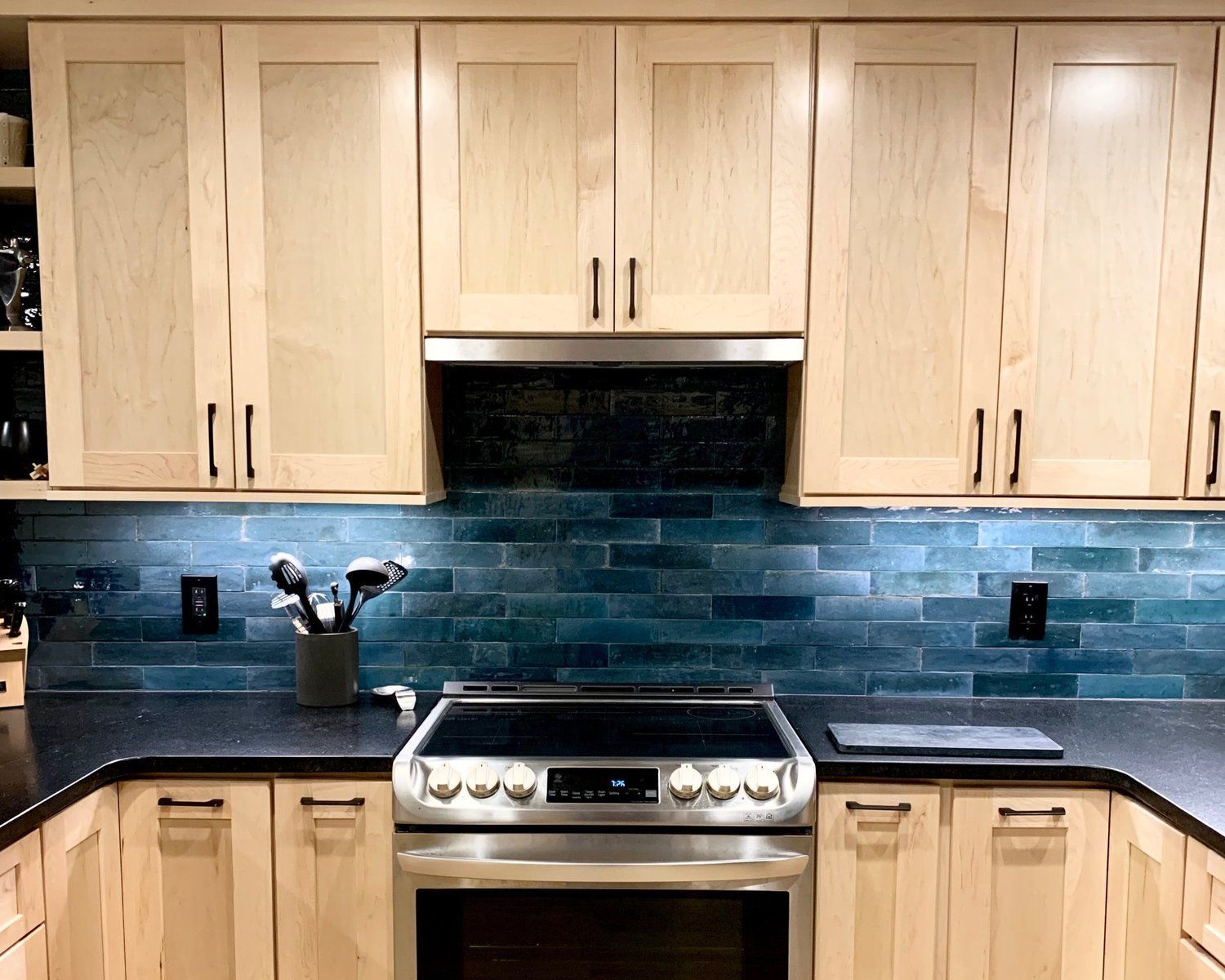 A kitchen stove with stainless steel appliances, light wood cabinets, and a glossy, dark teal subway tile backsplash.