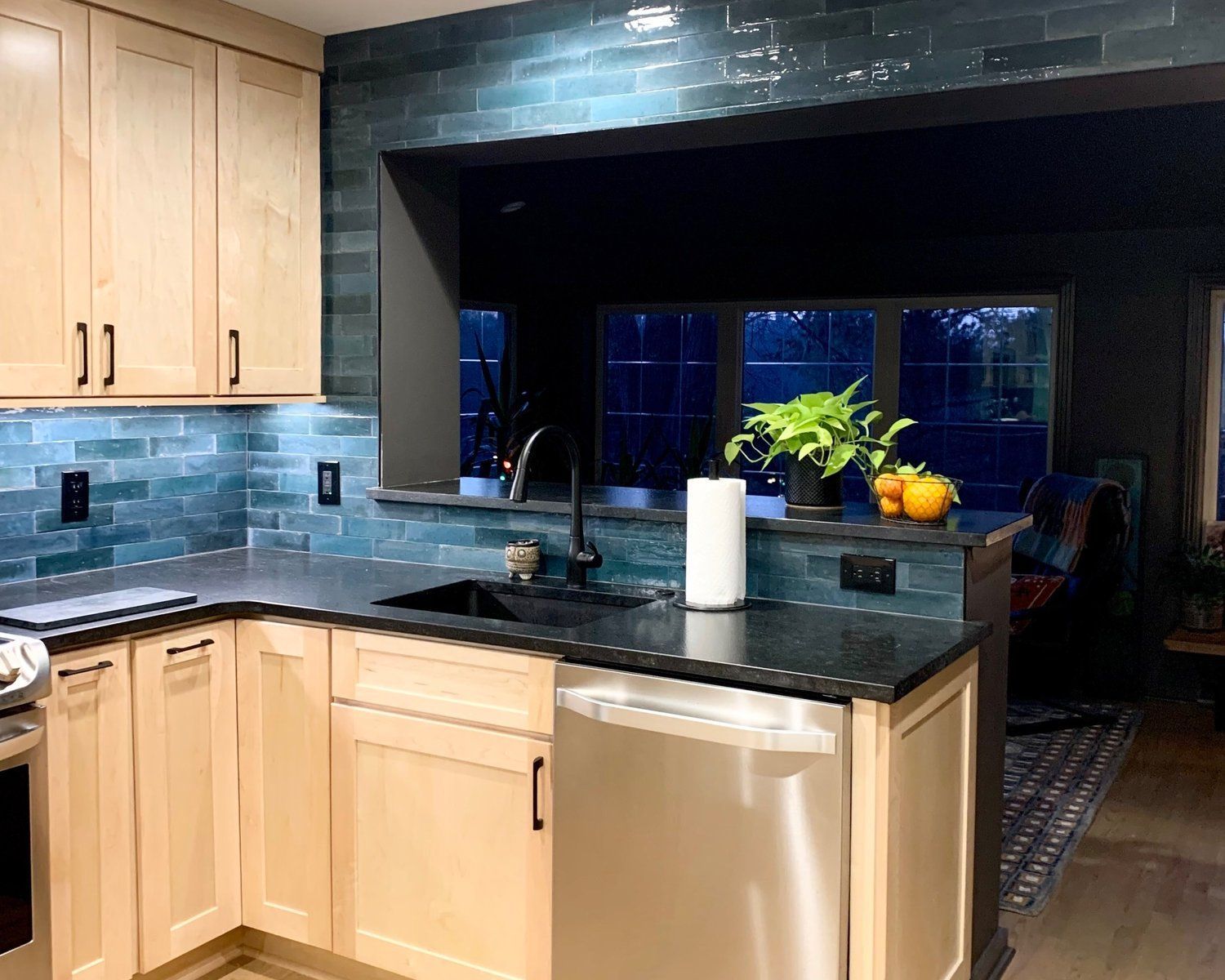 A kitchen with light wood cabinets, dark countertops, and a blue, glossy subway tile backsplash with a sink and window.