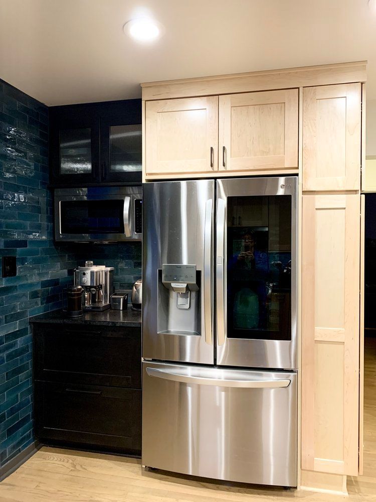 A modern kitchen with a stainless steel refrigerator flanked by light wood cabinetry against a dark blue tiled wall.