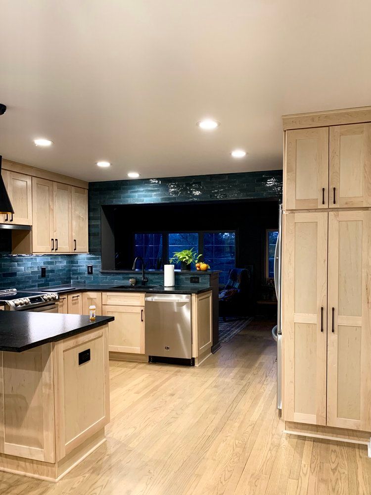 A modern kitchen with light wood cabinets, dark countertops, a stainless steel dishwasher, and a deep blue tile backsplash.
