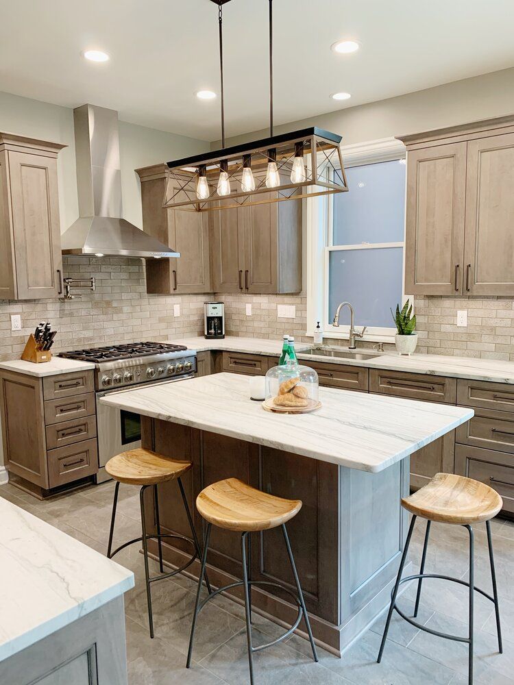 A modern kitchen features light wood cabinets, marble countertops, a large island with three stools, and a pendant light.
