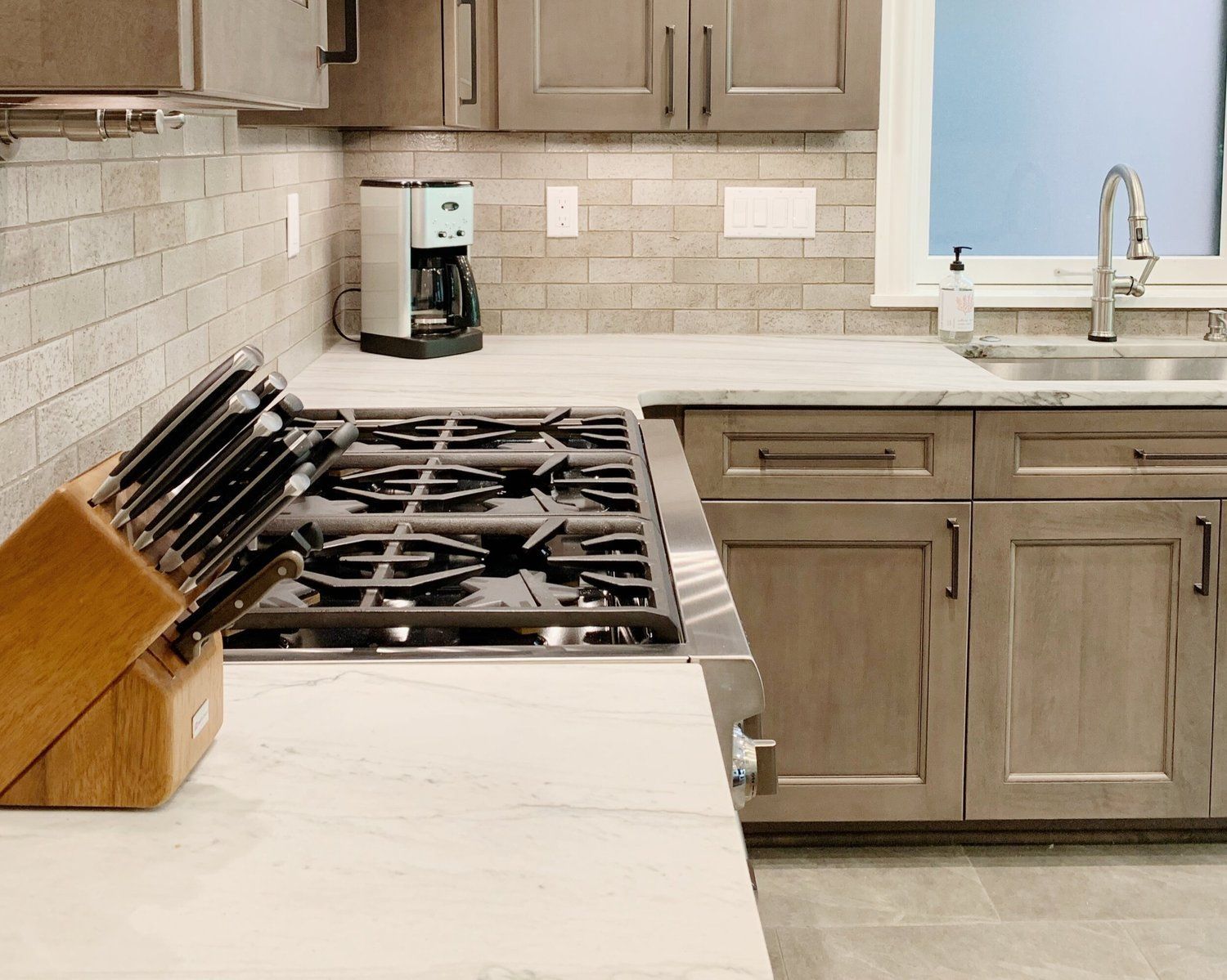 A kitchen countertop with a gas stove, a knife block, and gray cabinetry against a brick-style tiled backsplash.